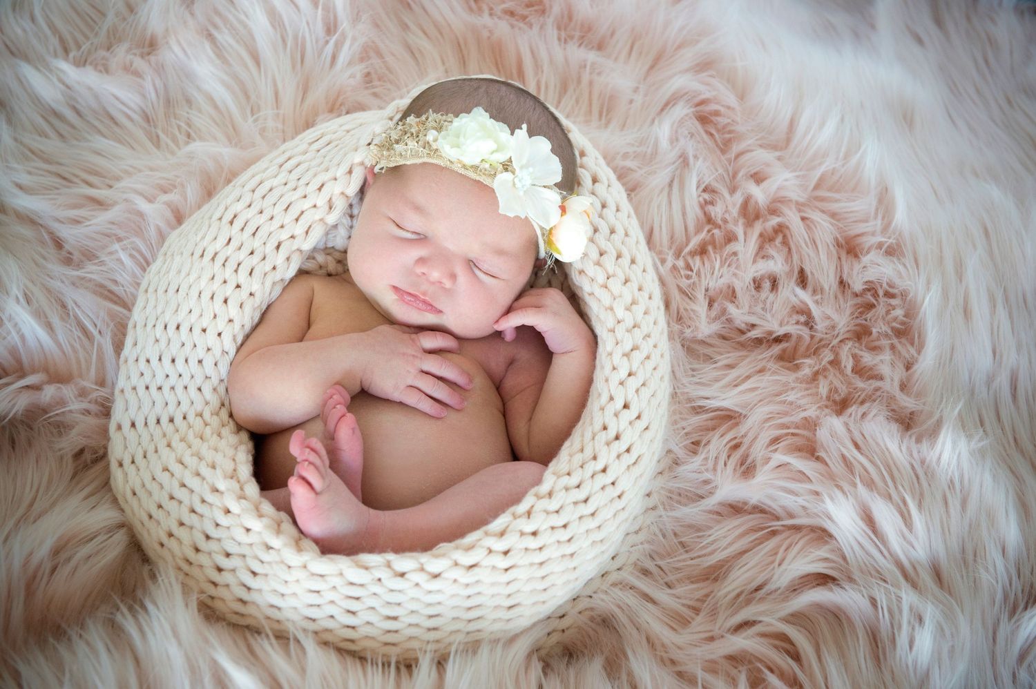 Peaceful newborn baby sleeping in a knit bowl with a floral headband on a soft pink fur blanket.