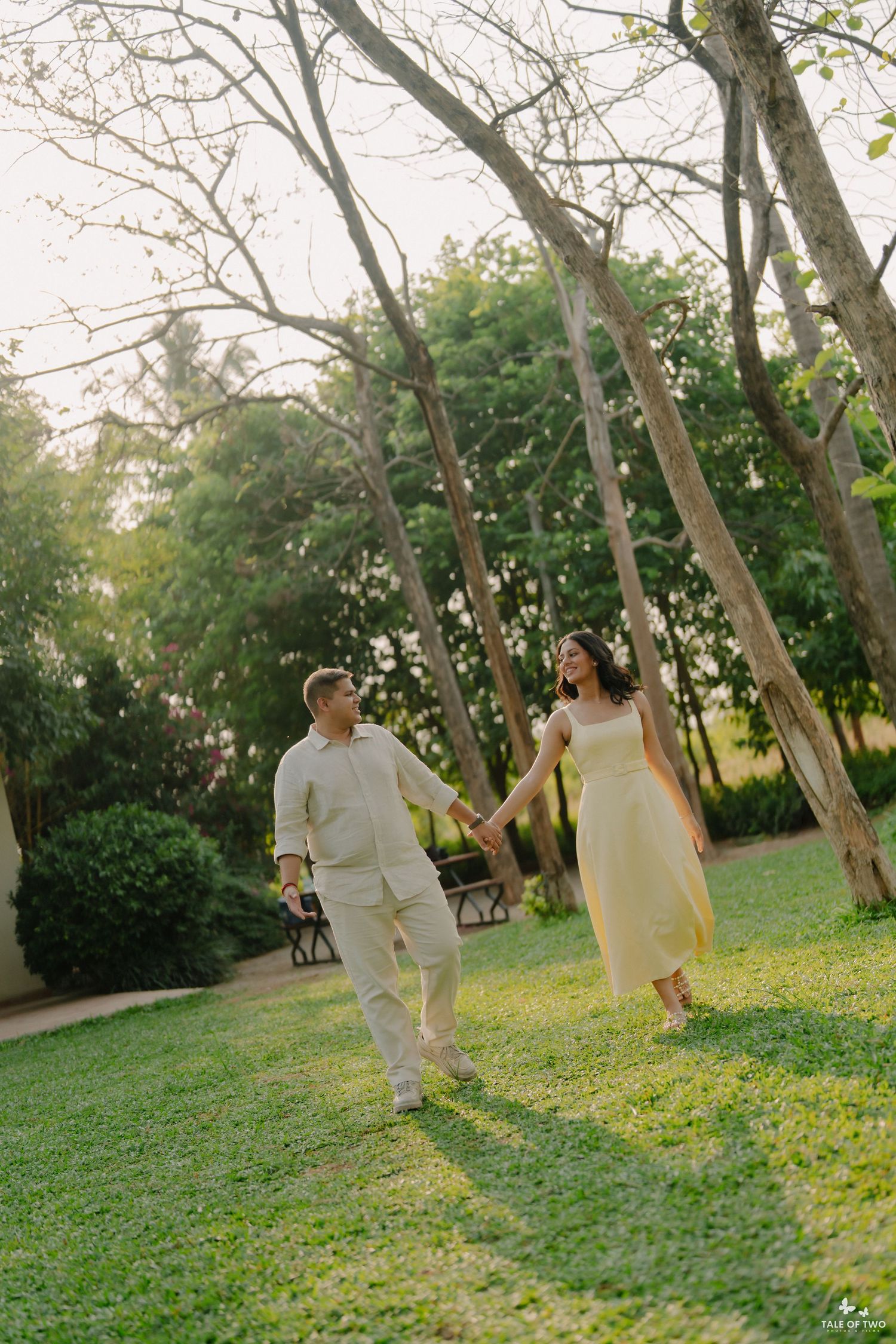 A couple in light outfits holds hands while walking through a lush green park surrounded by tall trees.
