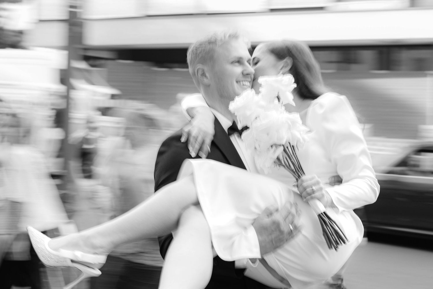 Groom carries bride in white dress and bouquet in black and white wedding photo with motion blur.