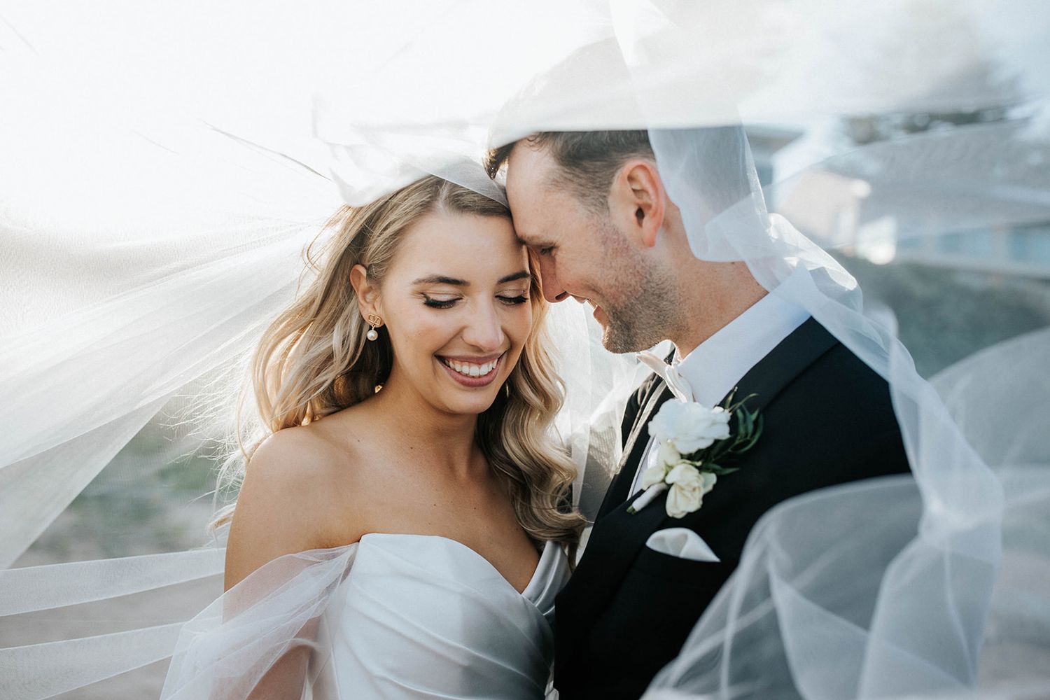 Bride and groom share an intimate moment under flowing veil, smiling together on their wedding day.
