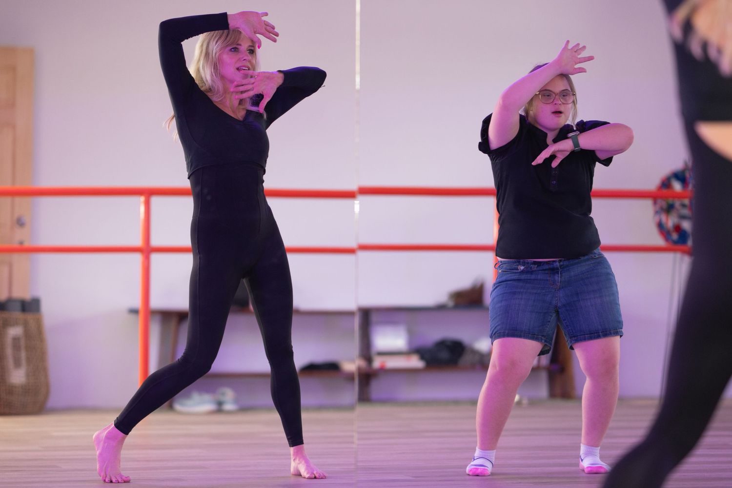 Two women in black outfits practice dance moves together in a pink-lit dance studio.
