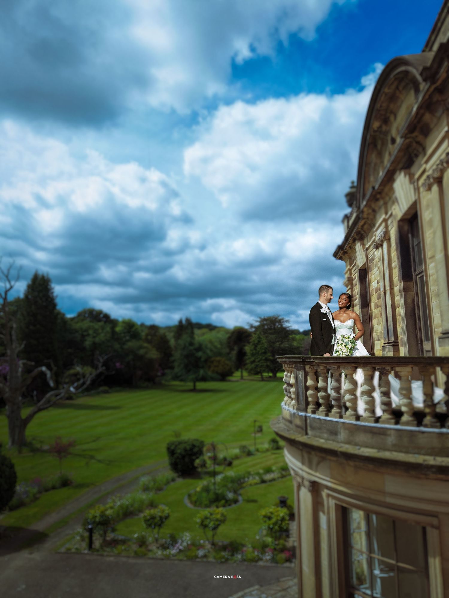 Bride and groom stand on ornate balcony of grand estate, overlooking lush green gardens under dramatic cloudy sky.