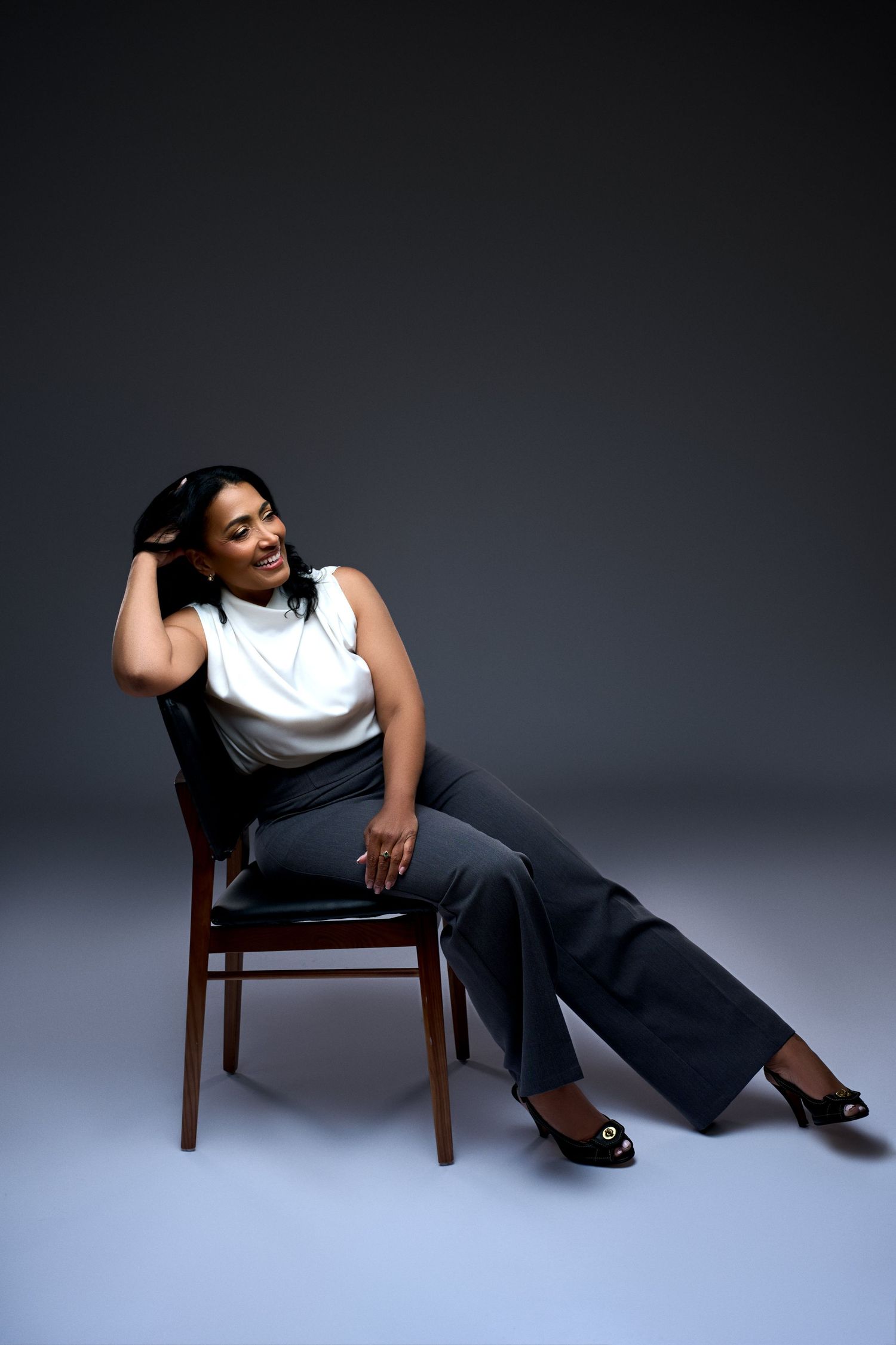 Woman in white top and gray wide-leg pants sits relaxed on a wooden chair, smiling, in a studio setting. New Jersey headshot photo