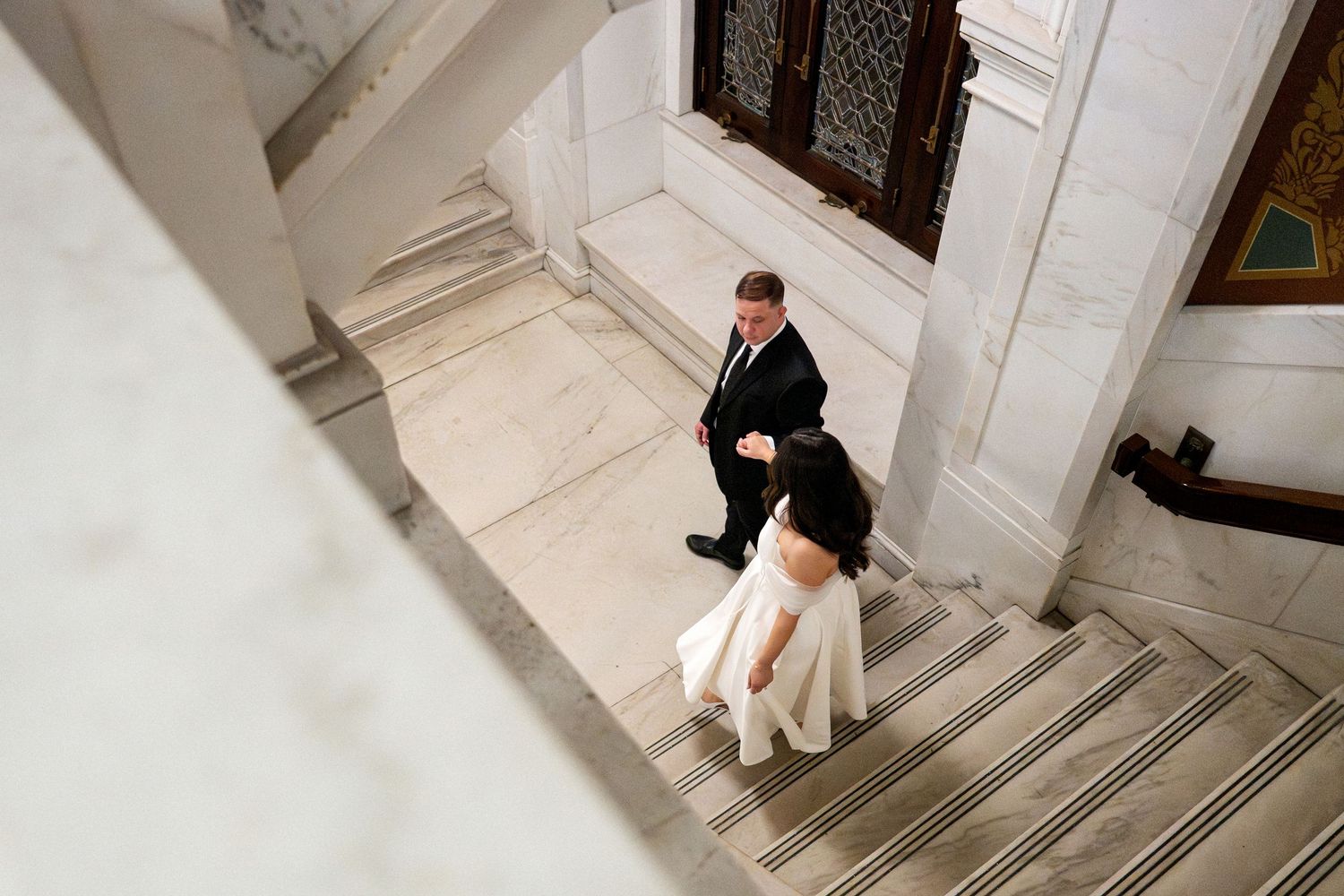 Bride and groom walking up marble stairs in elegant venue, aerial view of wedding couple in formal attire.