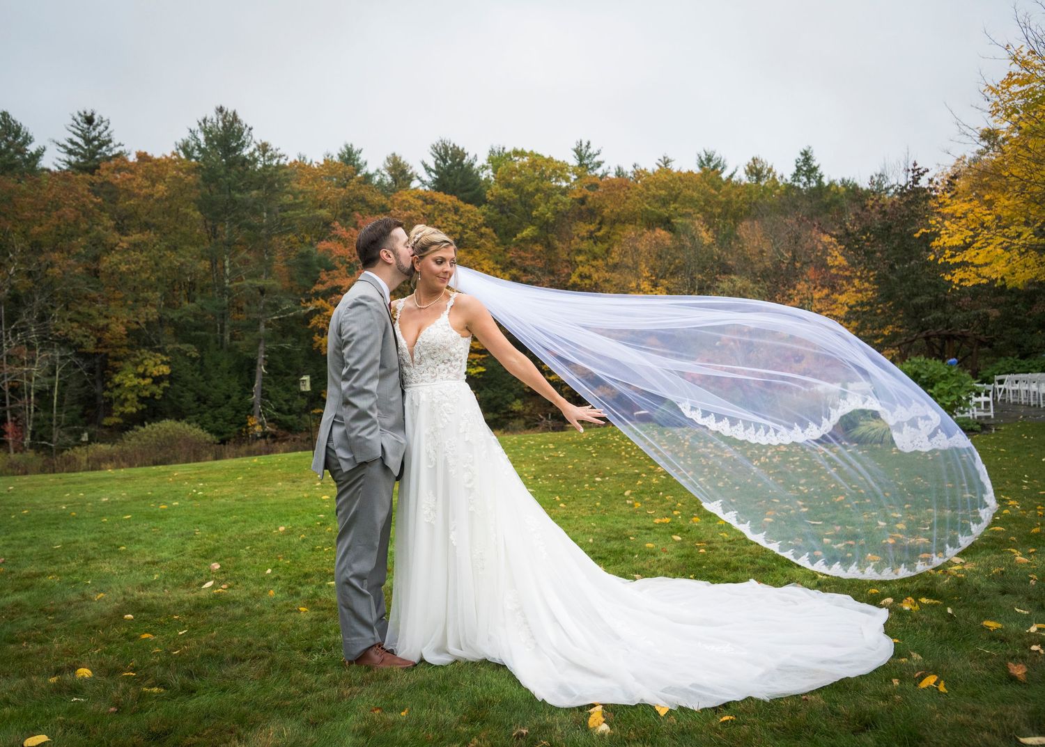 Bride and groom embrace on a grassy lawn as her long lace veil blows in the wind, with colorful autumn trees behind them.