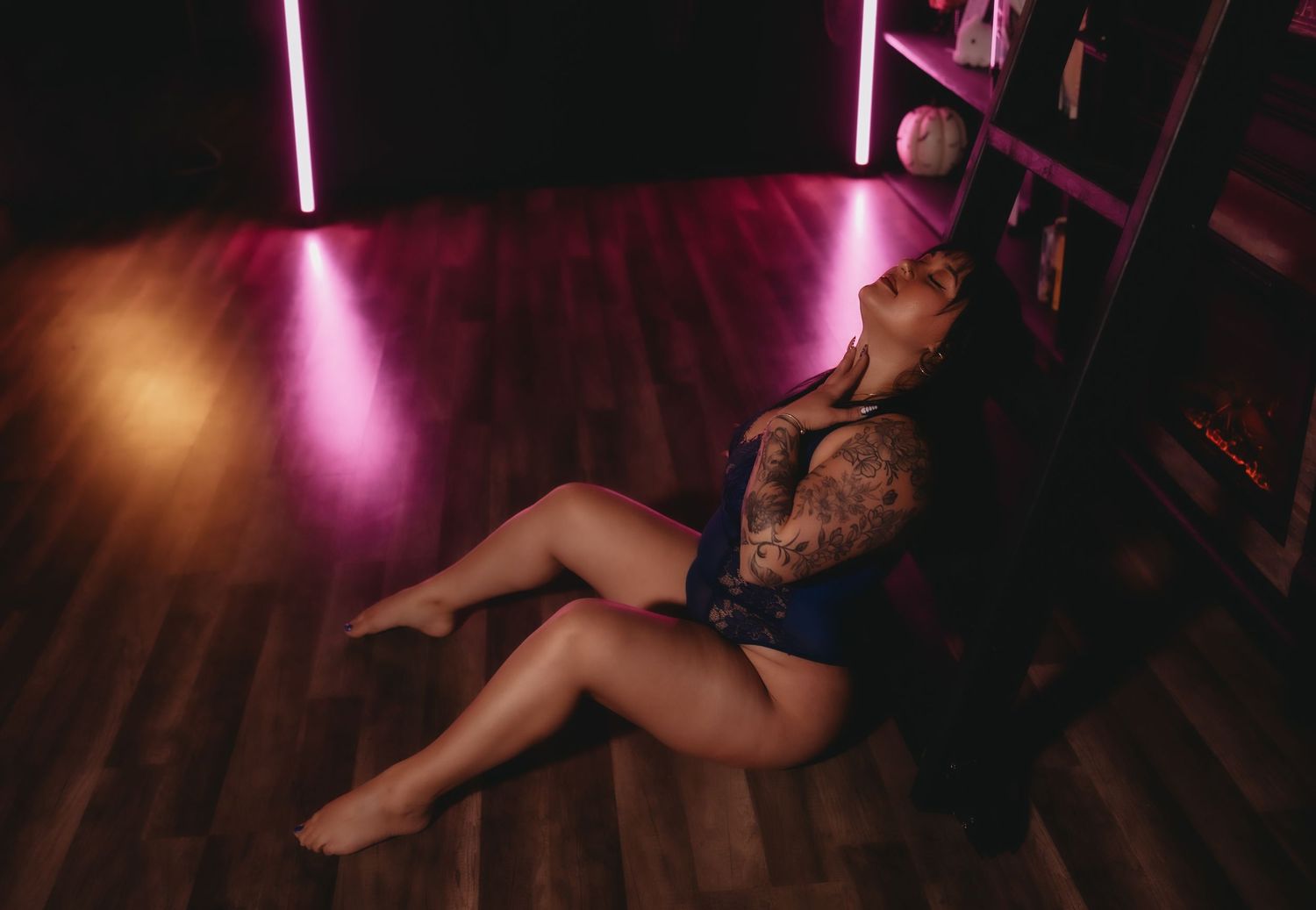 Woman with tattoos in blue outfit relaxing on hardwood floor beside bookshelf under pink neon light boudoir posing in brooksville florida