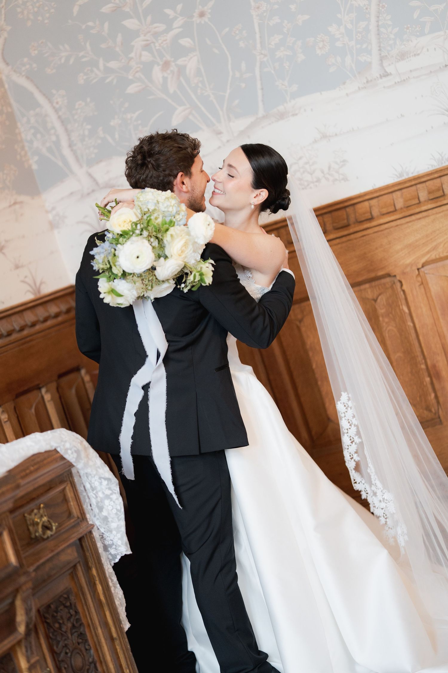 Bride and groom share a romantic embrace in elegant wedding attire, bride holding white floral bouquet with flowing veil.