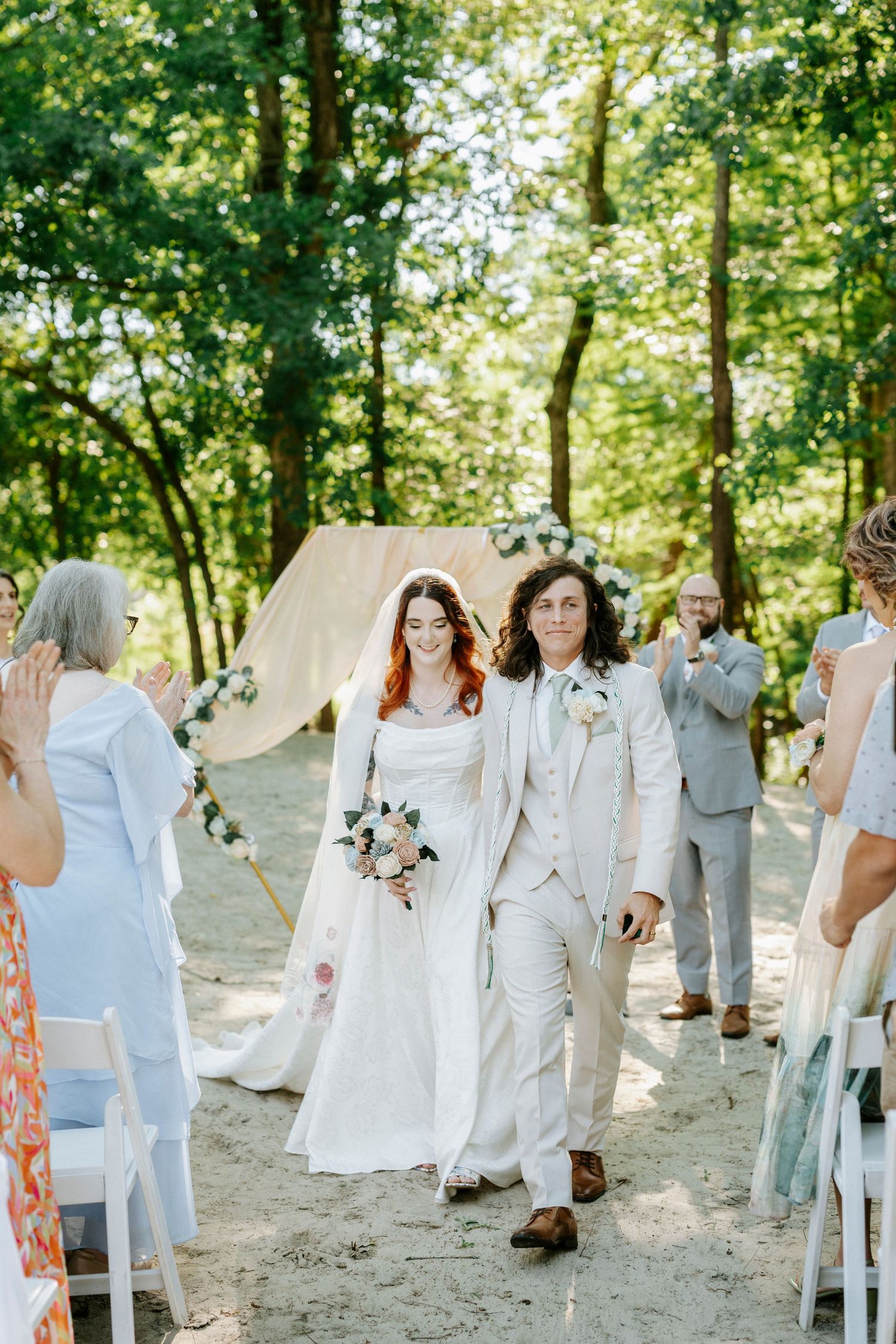 A bride and groom walk down the aisle at an outdoor woodland wedding ceremony, guests applauding as they exit.
