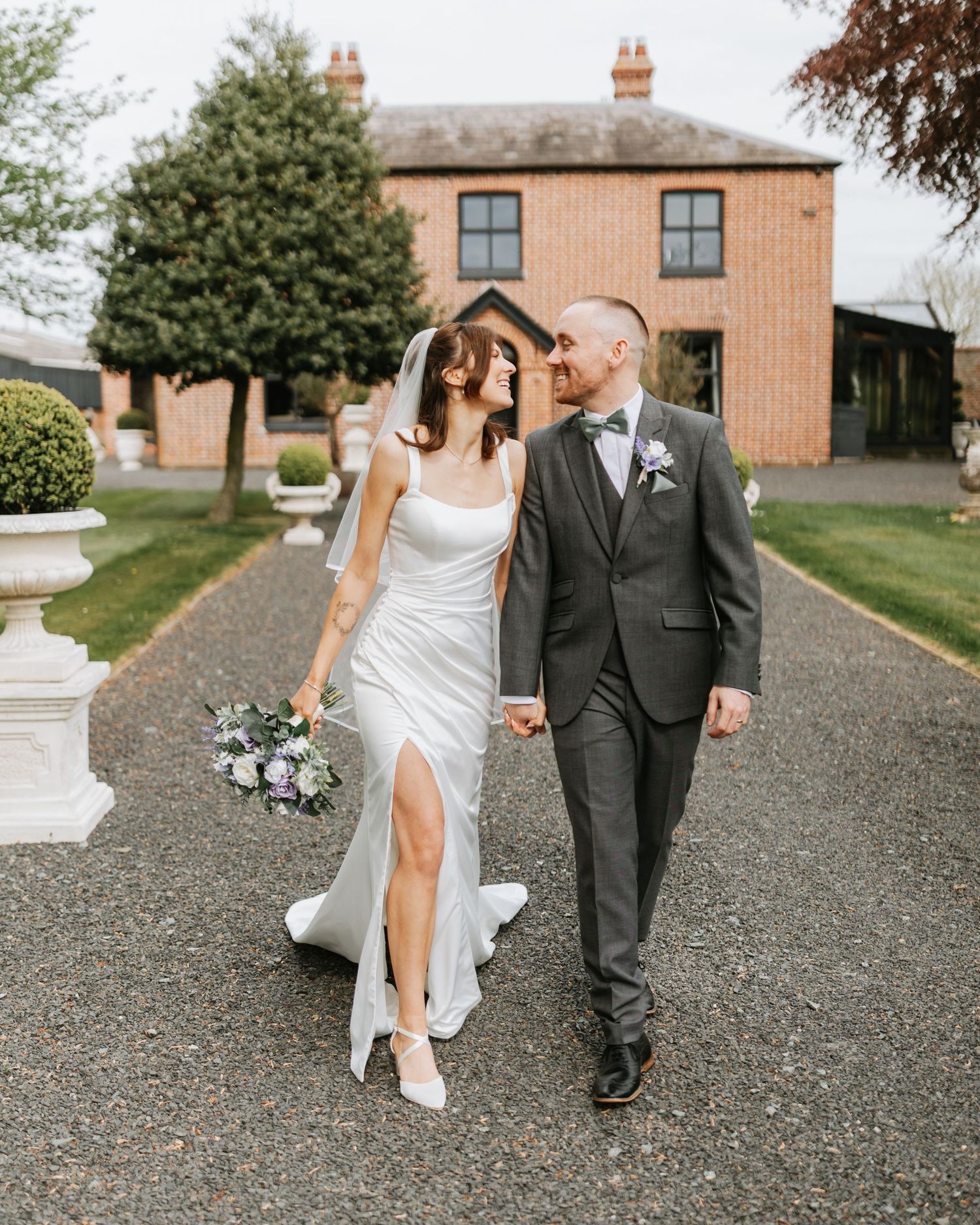 A bride and groom walk hand-in-hand on a gravel path outside a brick wedding venue, she in a white gown, he in a grey suit.