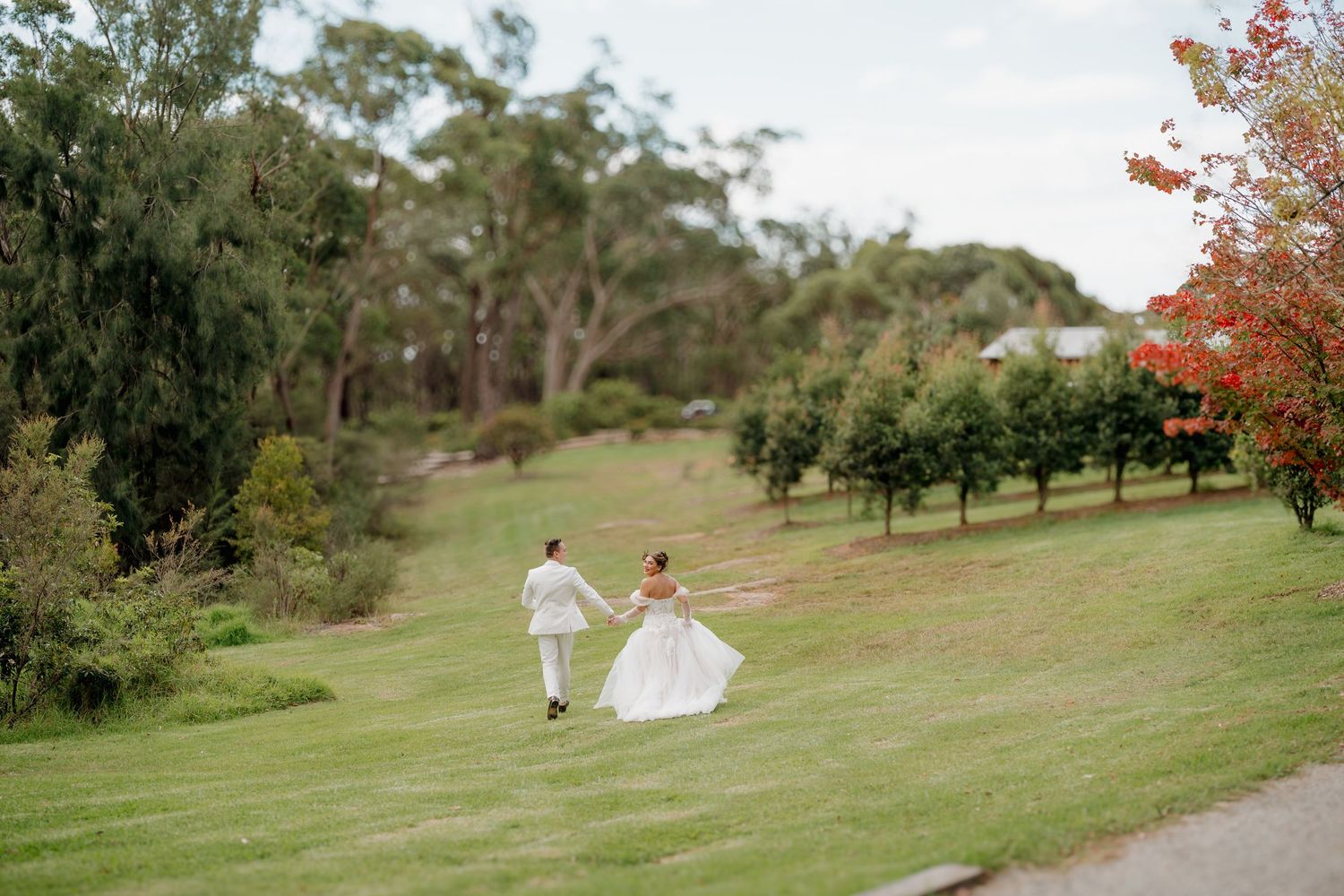 Bride and groom holding hands walking across lush green lawn surrounded by trees at outdoor wedding venue.