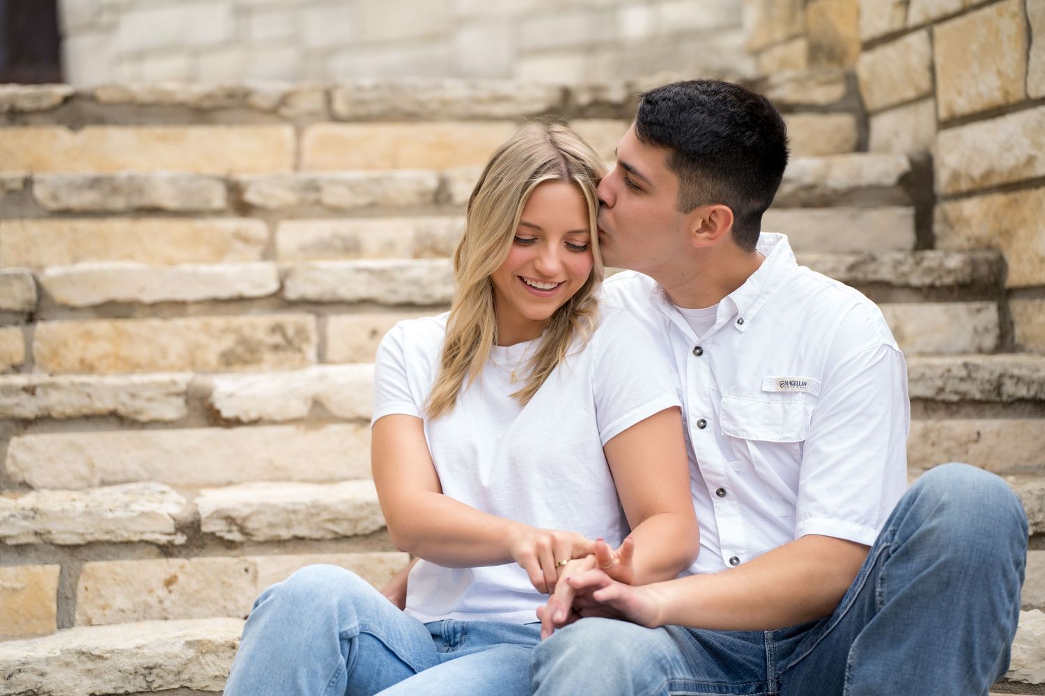 A couple sits on stone steps, sharing a tender kiss, both wearing white shirts and jeans in an outdoor setting.