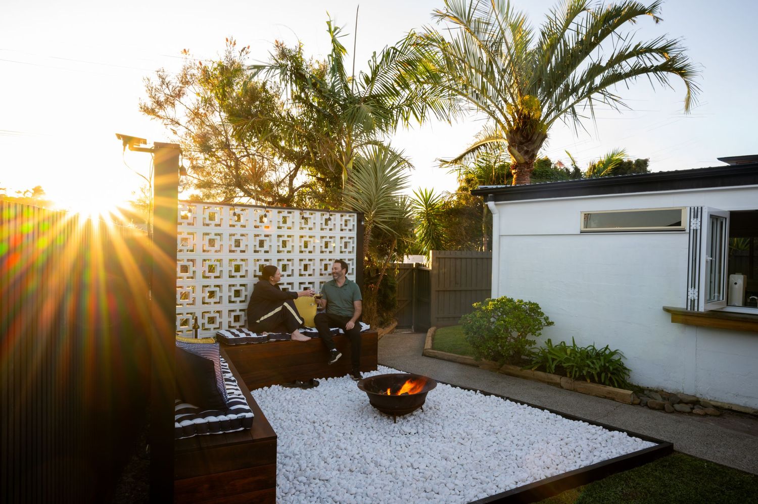 Two people relaxing in a modern outdoor patio with a fire pit, white gravel, and palm trees at sunset.