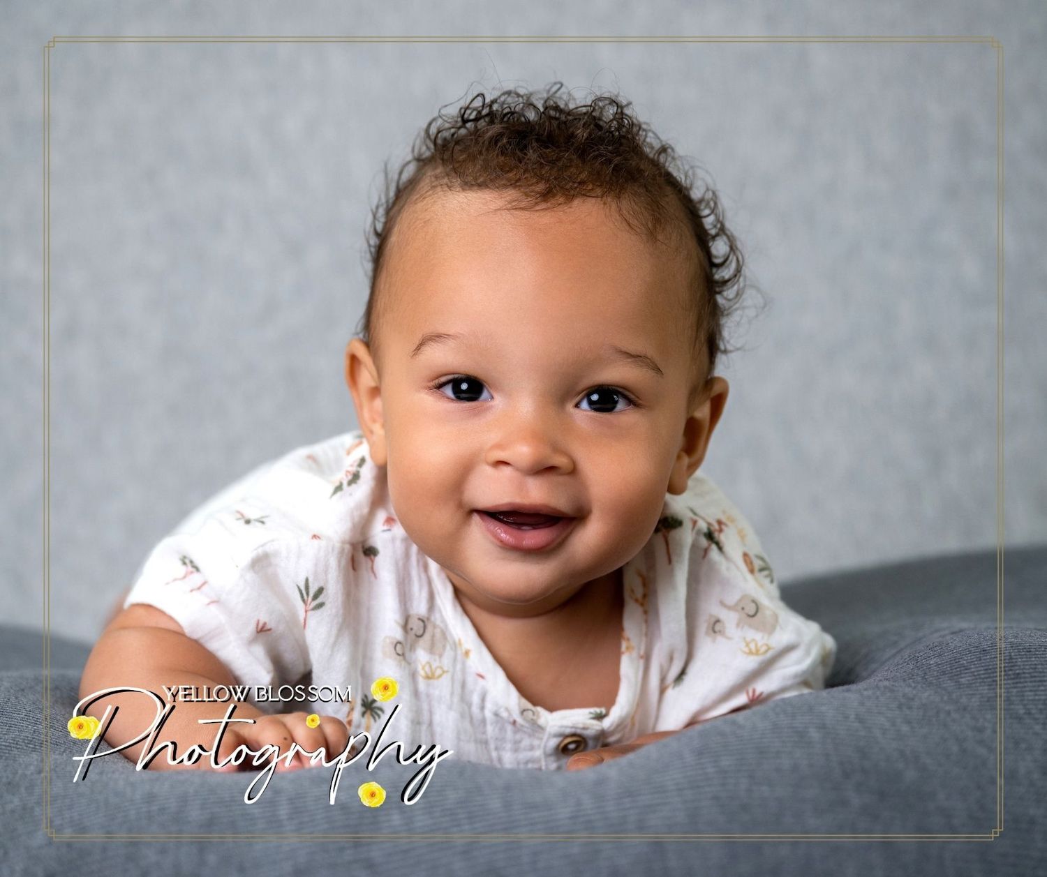 A happy baby wearing white floral clothing smiles while laying on a gray background during a portrait session.