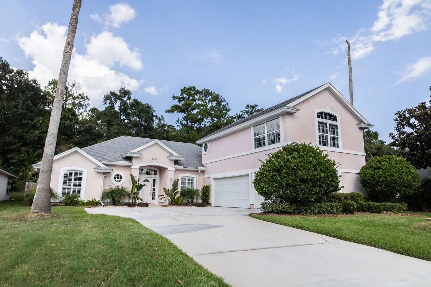 A large two-story house with white exterior and attached garage sits on a manicured lawn with palm trees.