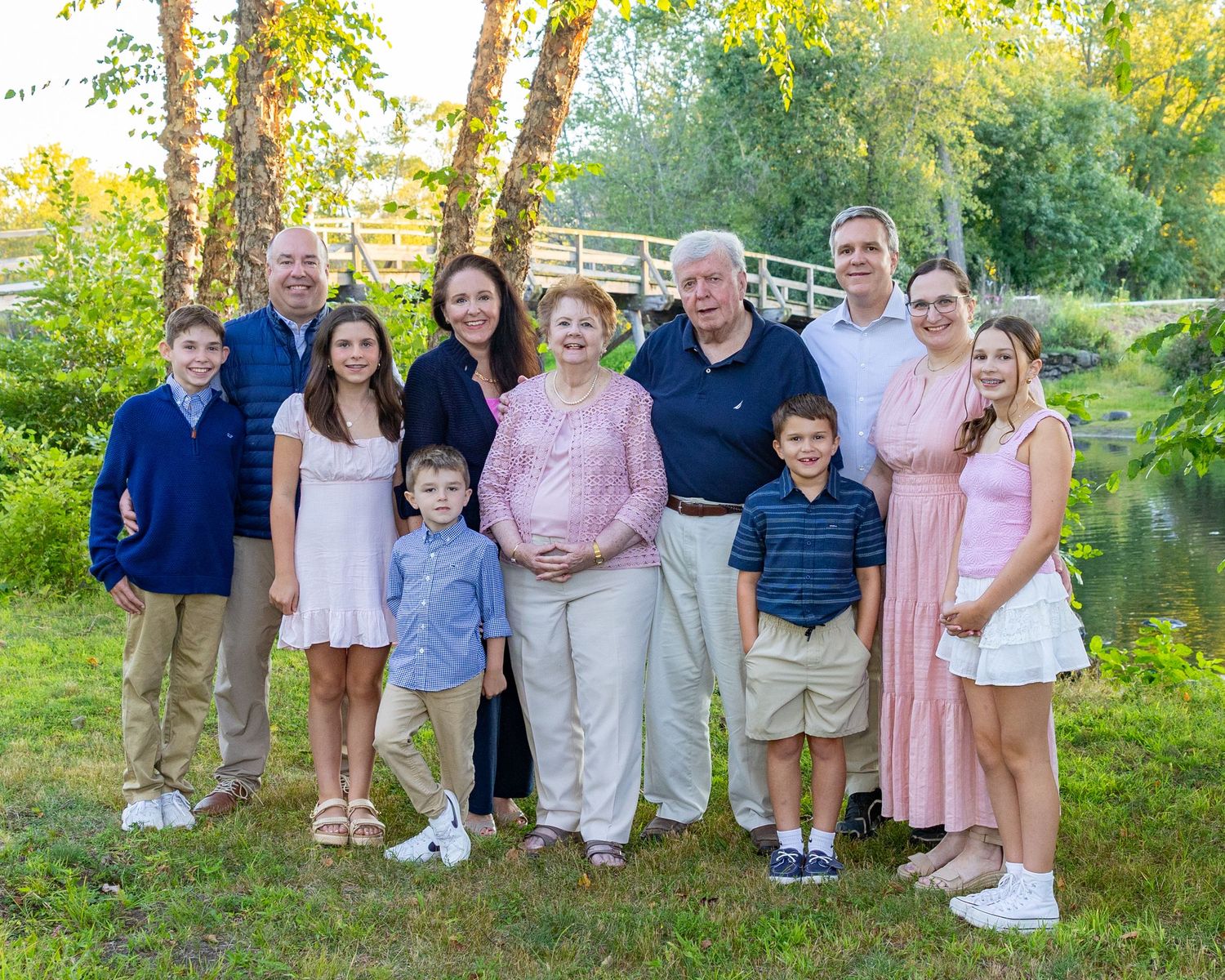A multi-generational family group poses together outdoors on a summer evening by Concord River with trees in the background.