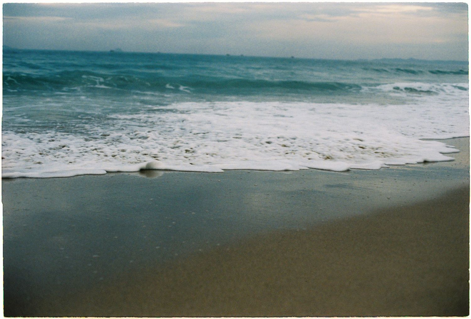 Gentle ocean waves washing onto a sandy beach under a soft cloudy sky at dusk.