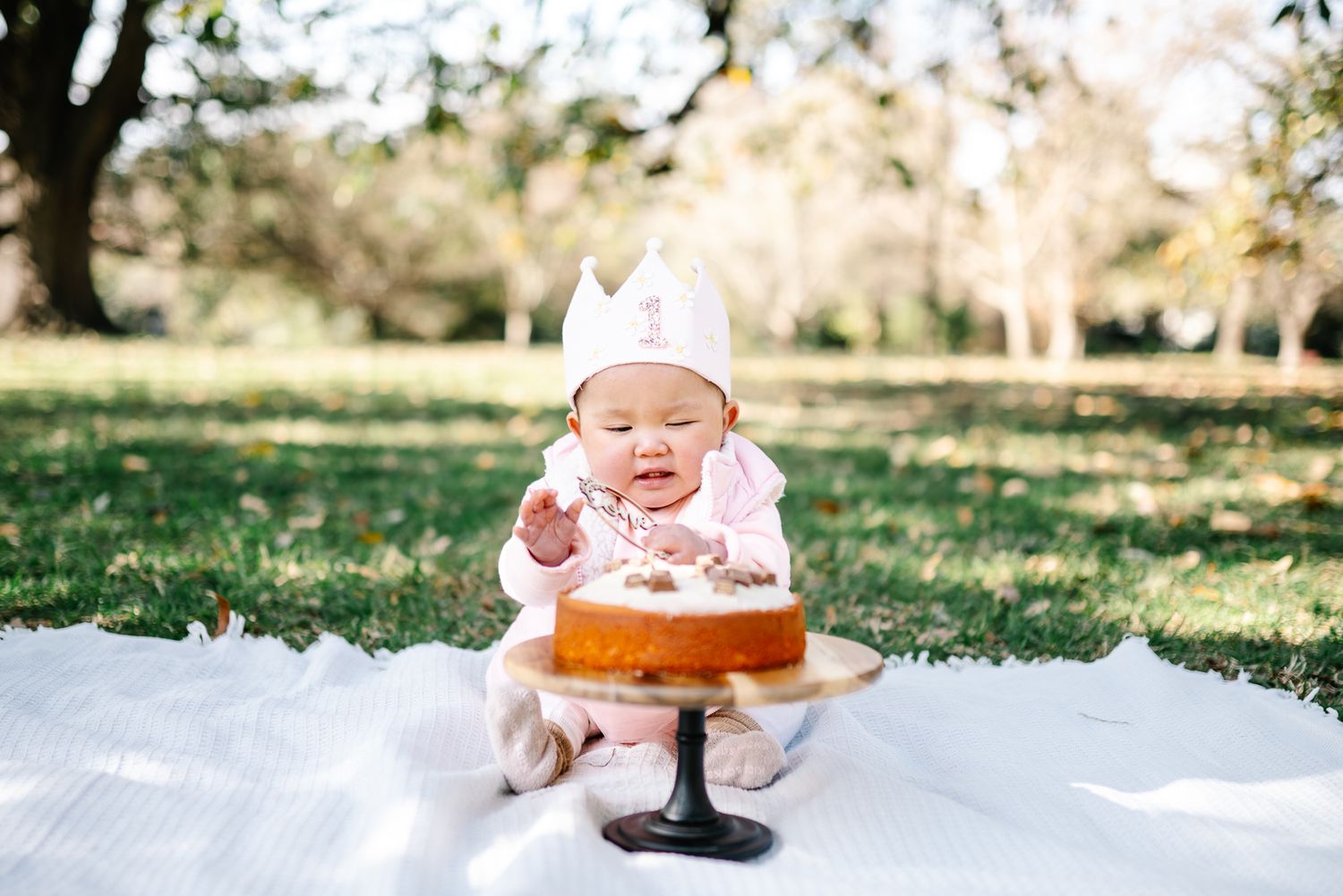 Baby sits on white blanket in a Melbourne park celebrating her first birthday with a cake smash photography.