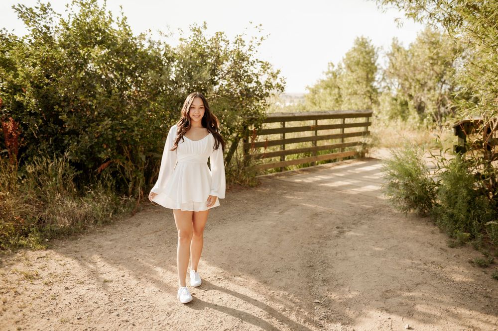 A figure in a flowy white dress and sneakers walks along a sunlit dirt path bordered by wooden fencing and greenery.