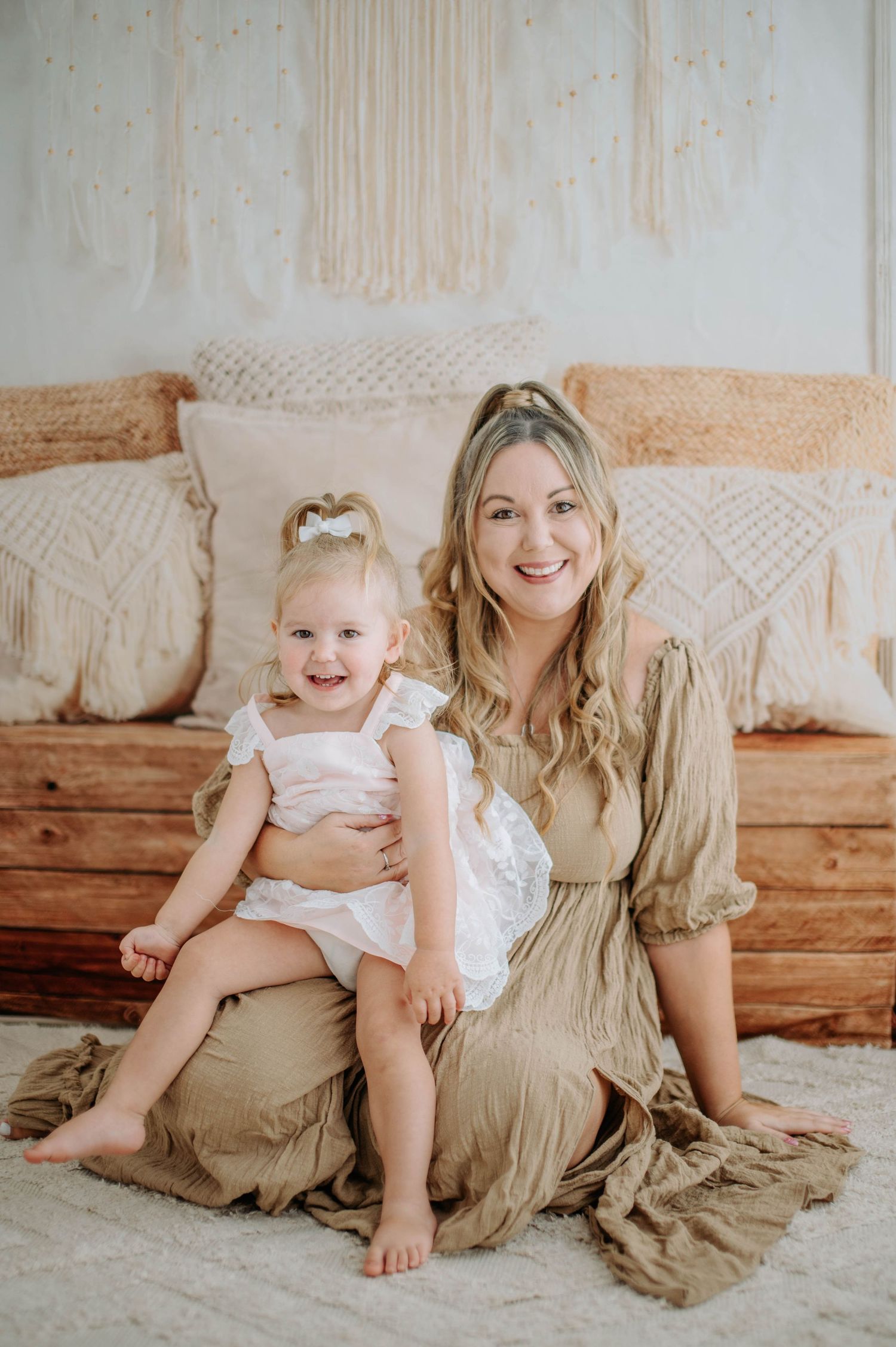 A small child wearing a white lace dress sits on a couch with decorative macrame pillows.