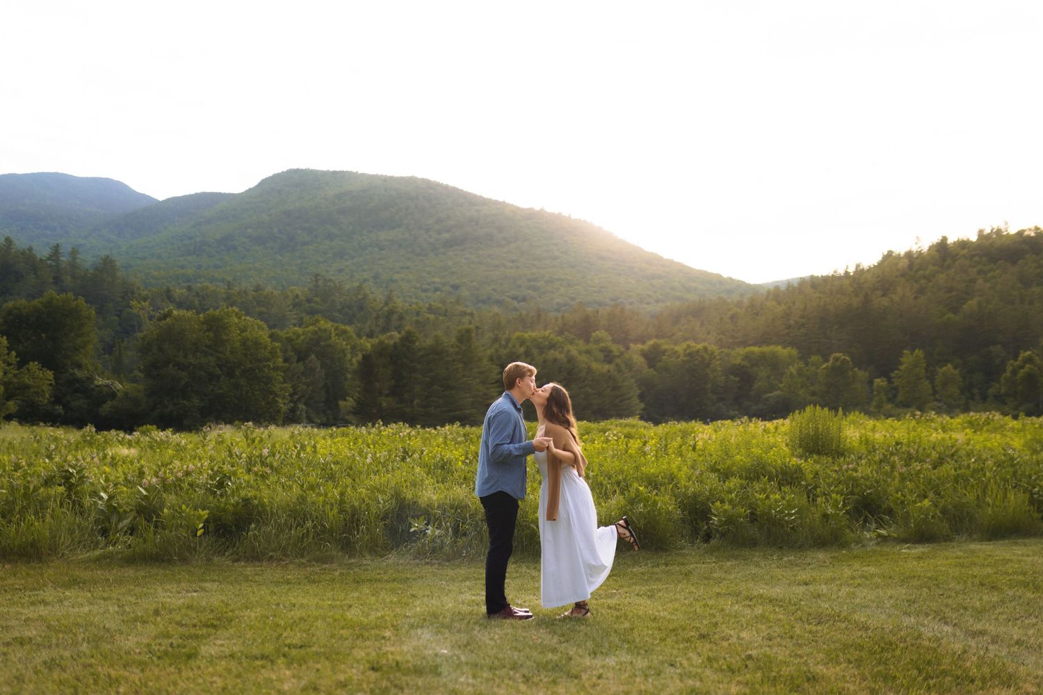 A couple embraces in a scenic mountain meadow at golden hour with rolling hills and forests in the background.