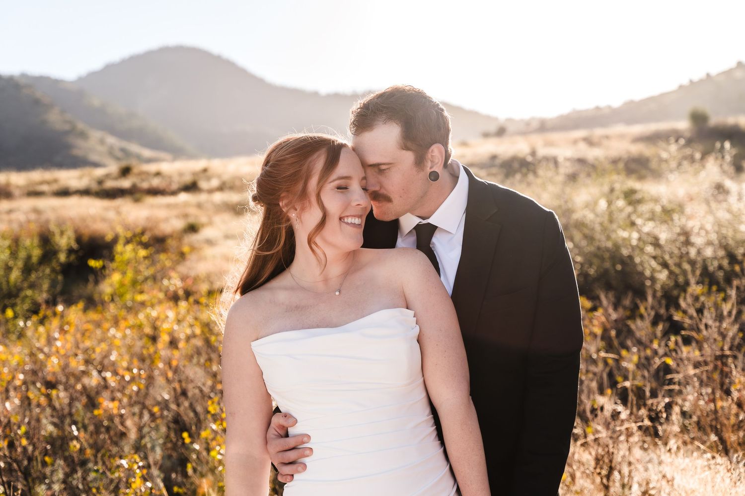 Newlywed couple shares romantic moments in golden sunlit field with mountain backdrop during sunset photoshoot.