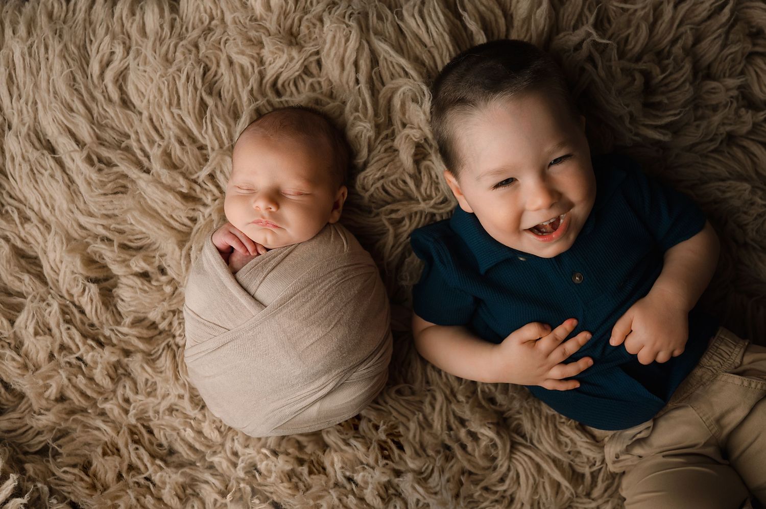 Two children lying on a beige fluffy rug, one swaddled in cloth while the other wears a navy shirt and smiles.
