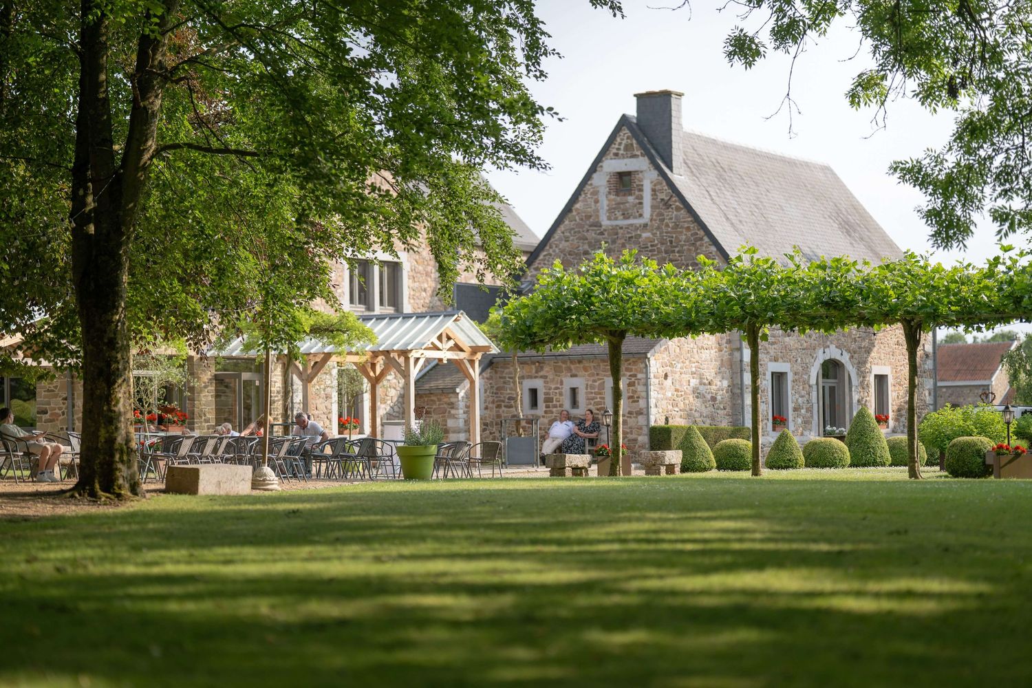 Elegant stone country house with wrap-around porch and manicured lawn surrounded by green trees on a sunny day.