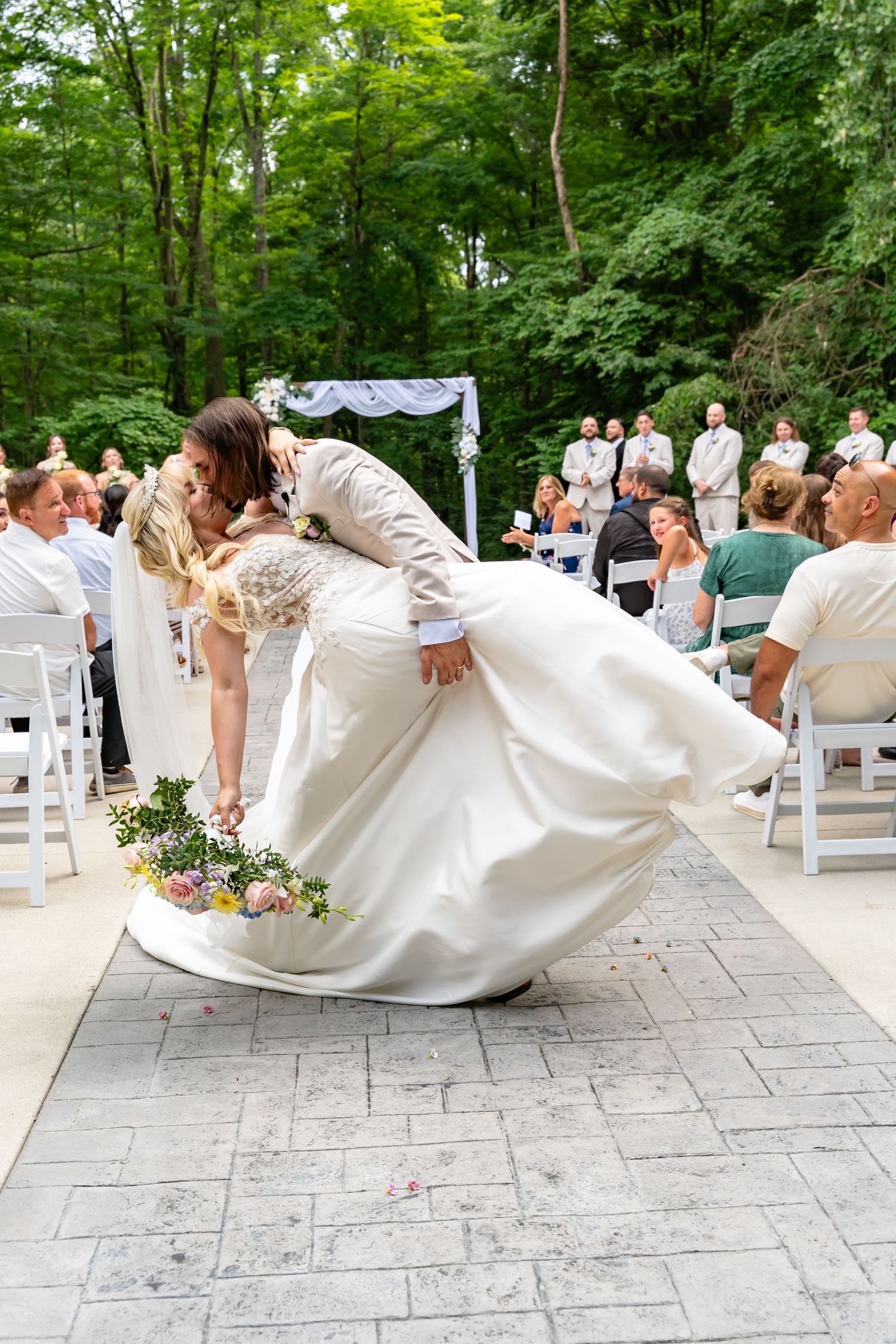 A romantic outdoor wedding ceremony dip pose on a stone patio surrounded by lush green trees.