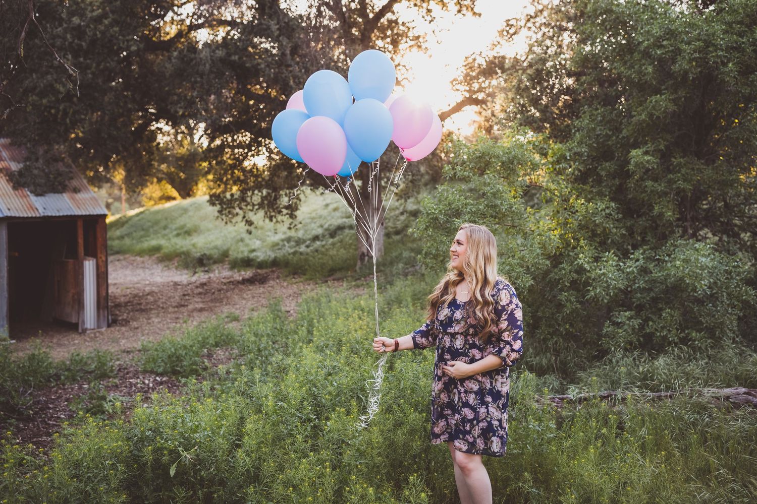 A person in a floral dress holds colorful balloons in a scenic outdoor setting during golden hour.