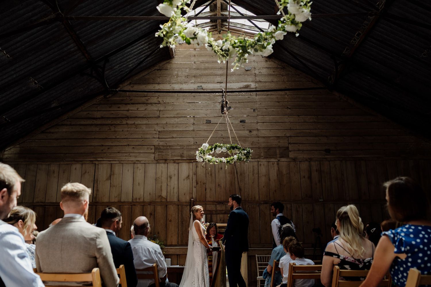 Wedding ceremony setup in rustic wooden barn with white floral arrangements and natural lighting.