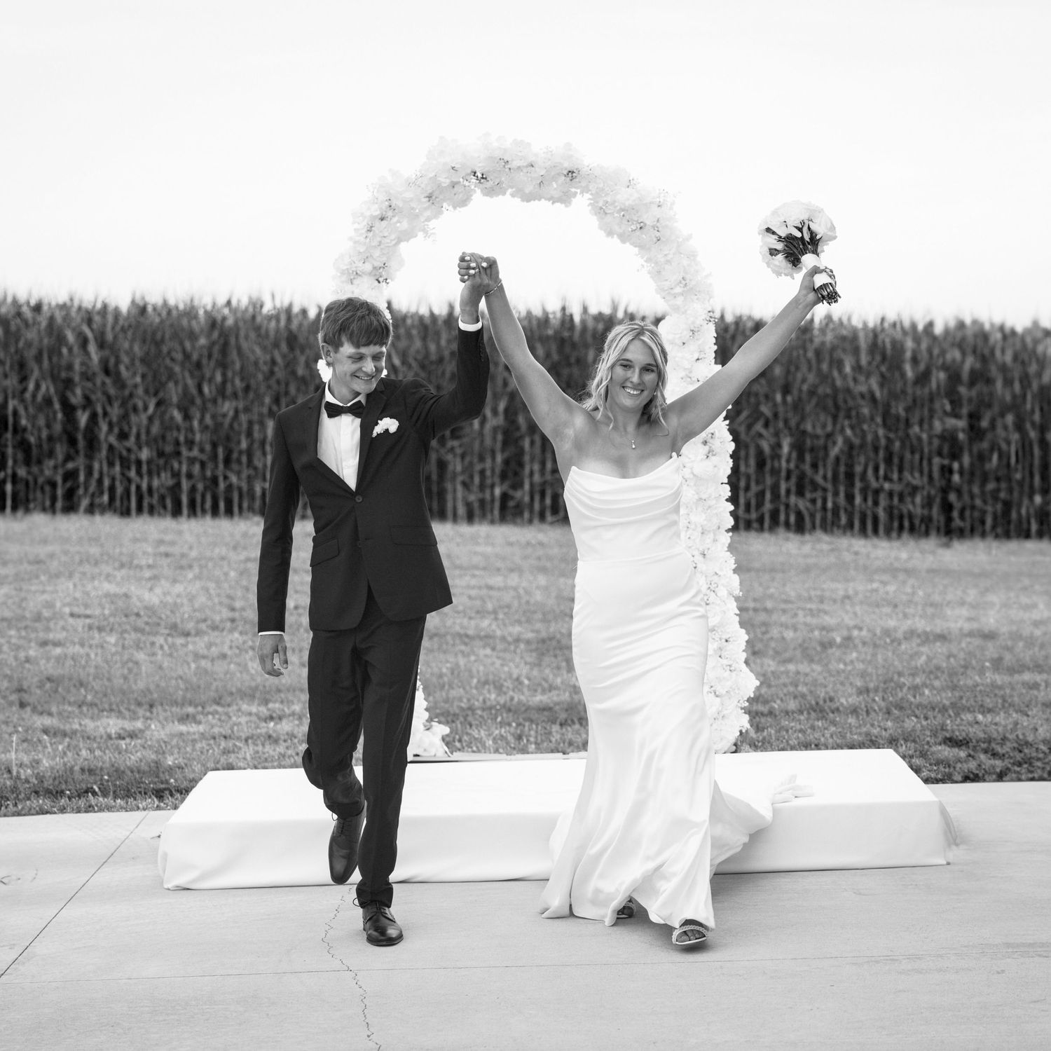 Joyful couple celebrates at outdoor wedding ceremony with floral arch and cornfield backdrop in black and white.