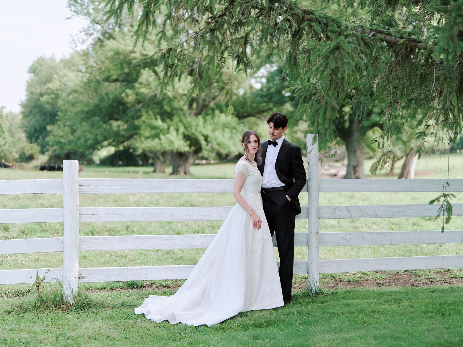 A wedding couple poses together outdoors, with the bride in a lace gown and groom wearing a yellow tie.