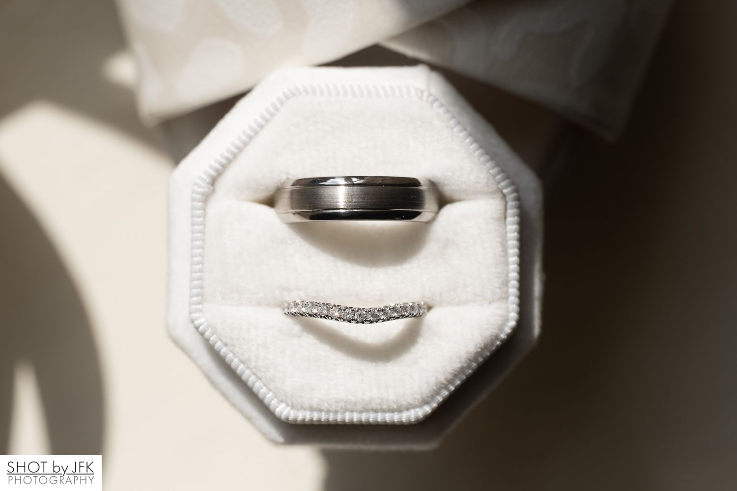 A silver wedding band sits in a white octagonal ring box casting dramatic shadows.