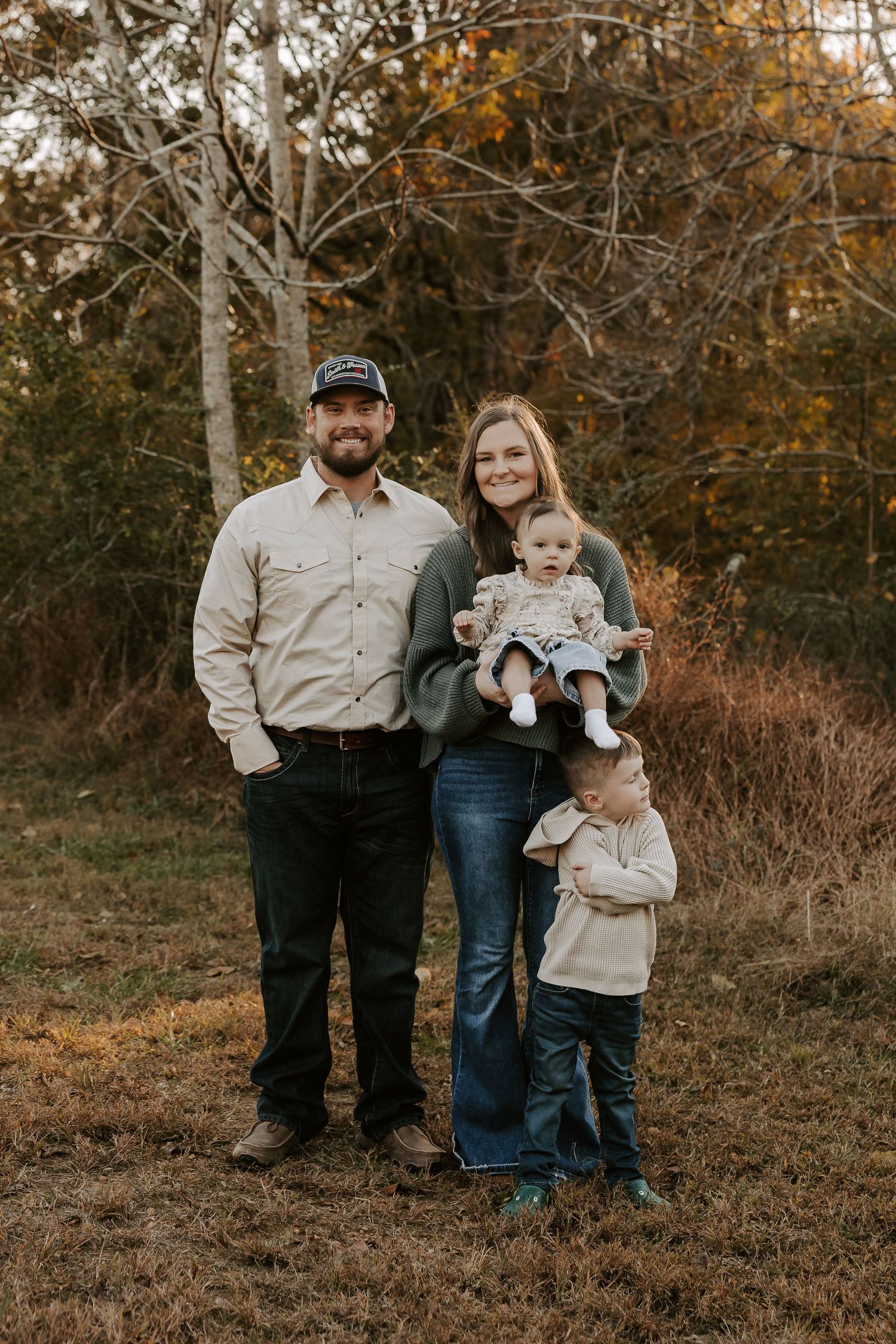 A family in coordinating beige outfits poses together on a grassy hillside during golden autumn sunset.
