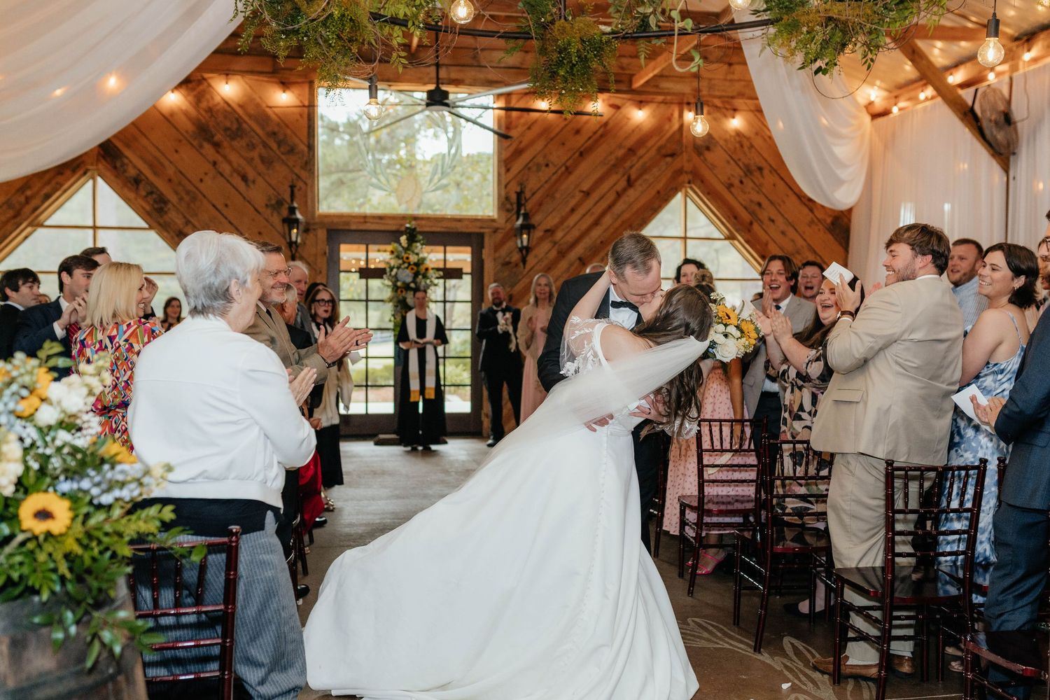 Newlyweds share their first dance surrounded by guests in a rustic barn venue with twinkling lights and wooden beams.