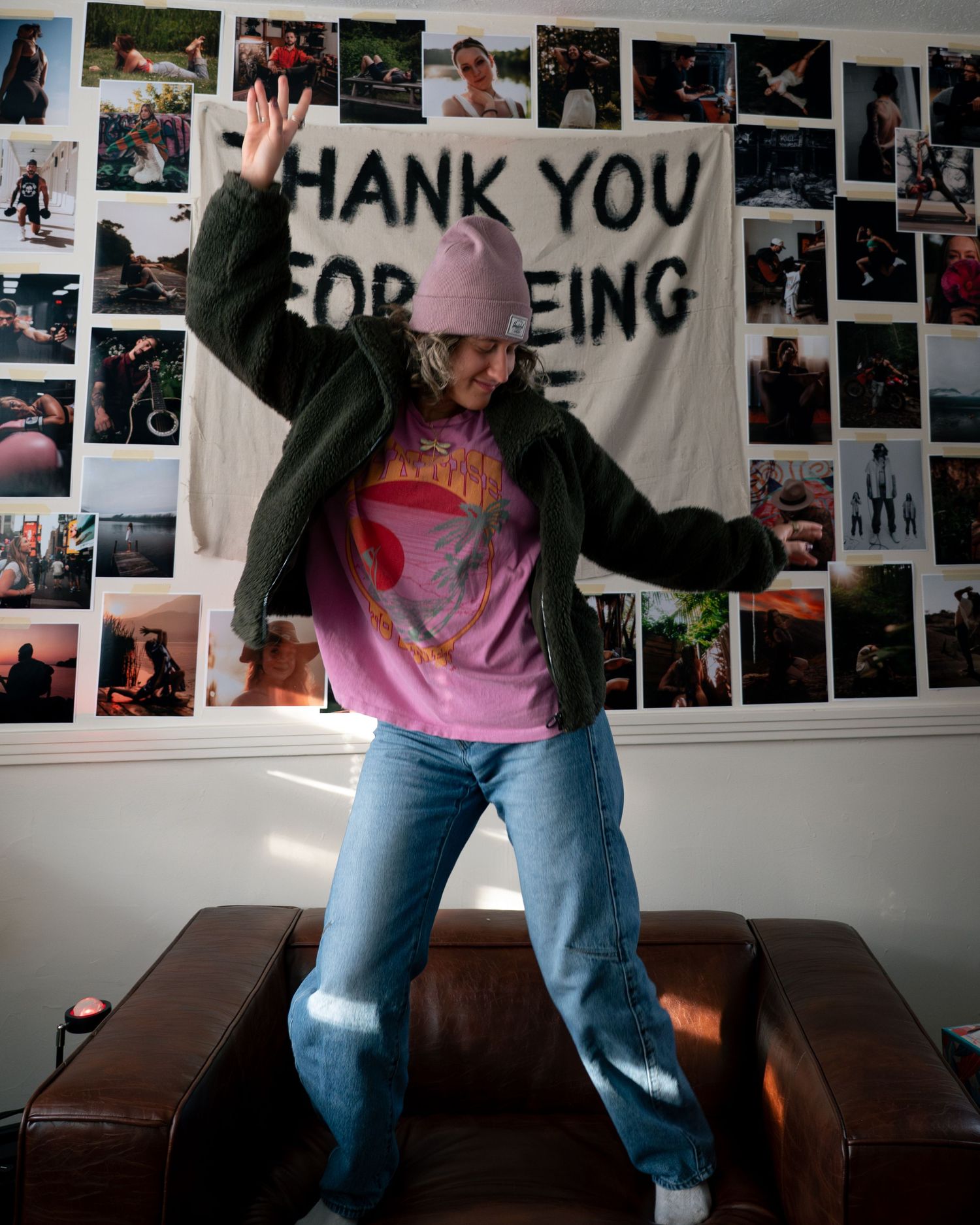 Someone dances energetically in front of a wall covered with photos and a banner that says 'THANK YOU FOR BEING'.