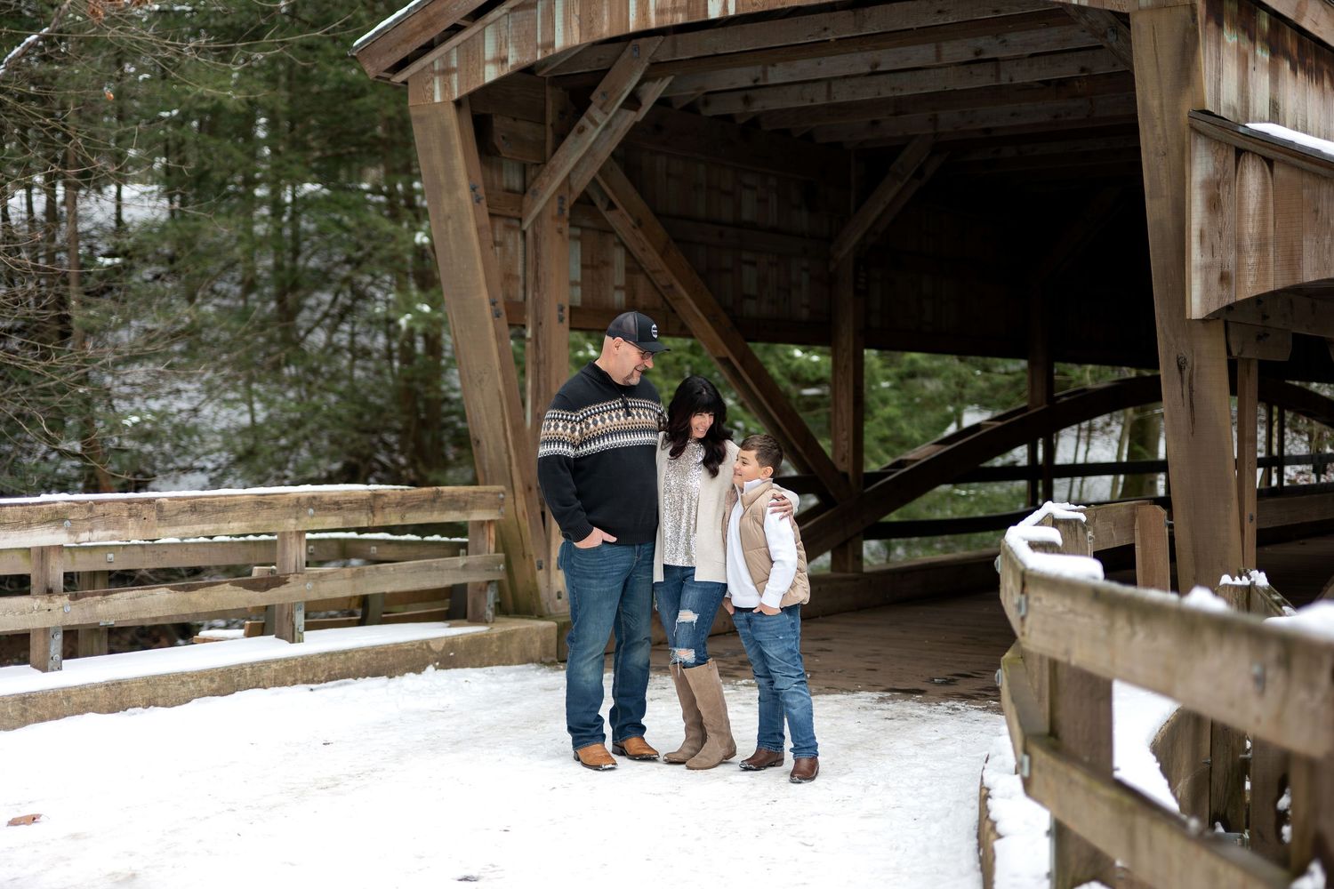 Family of three standing on a snowy covered bridge surrounded by winter scenery.