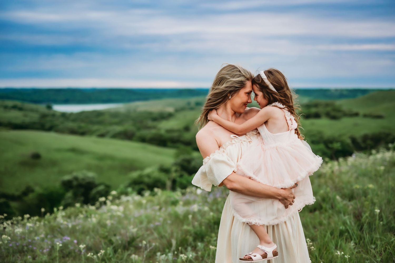 Couple sharing an intimate embrace in a romantic hillside field with blooming wildflowers under a blue sky.