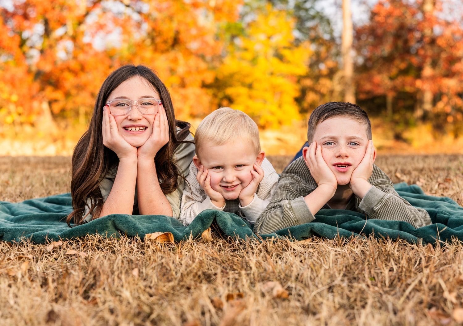 Four people lying in autumn leaves with bright orange and yellow fall foliage in the background.