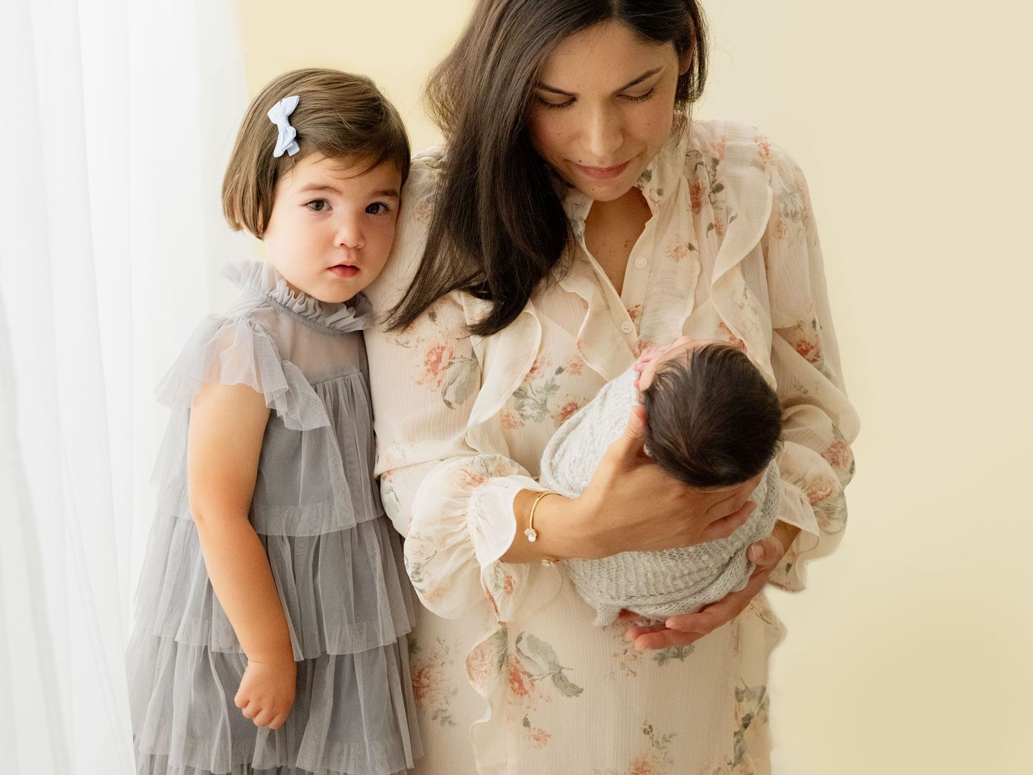 A toddler in gray dress gazes at tender moment between parent and baby in a soft natural light setting.