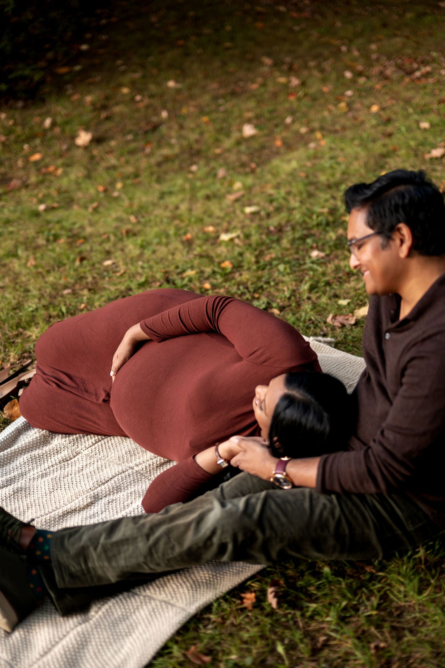 Pregnant woman resting head on partner's lap on blanket in green autumn park setting.