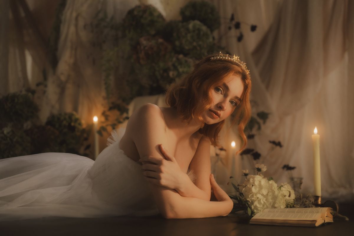 A bride in a flowing white wedding dress stands gracefully against sheer curtains in a softly lit room.