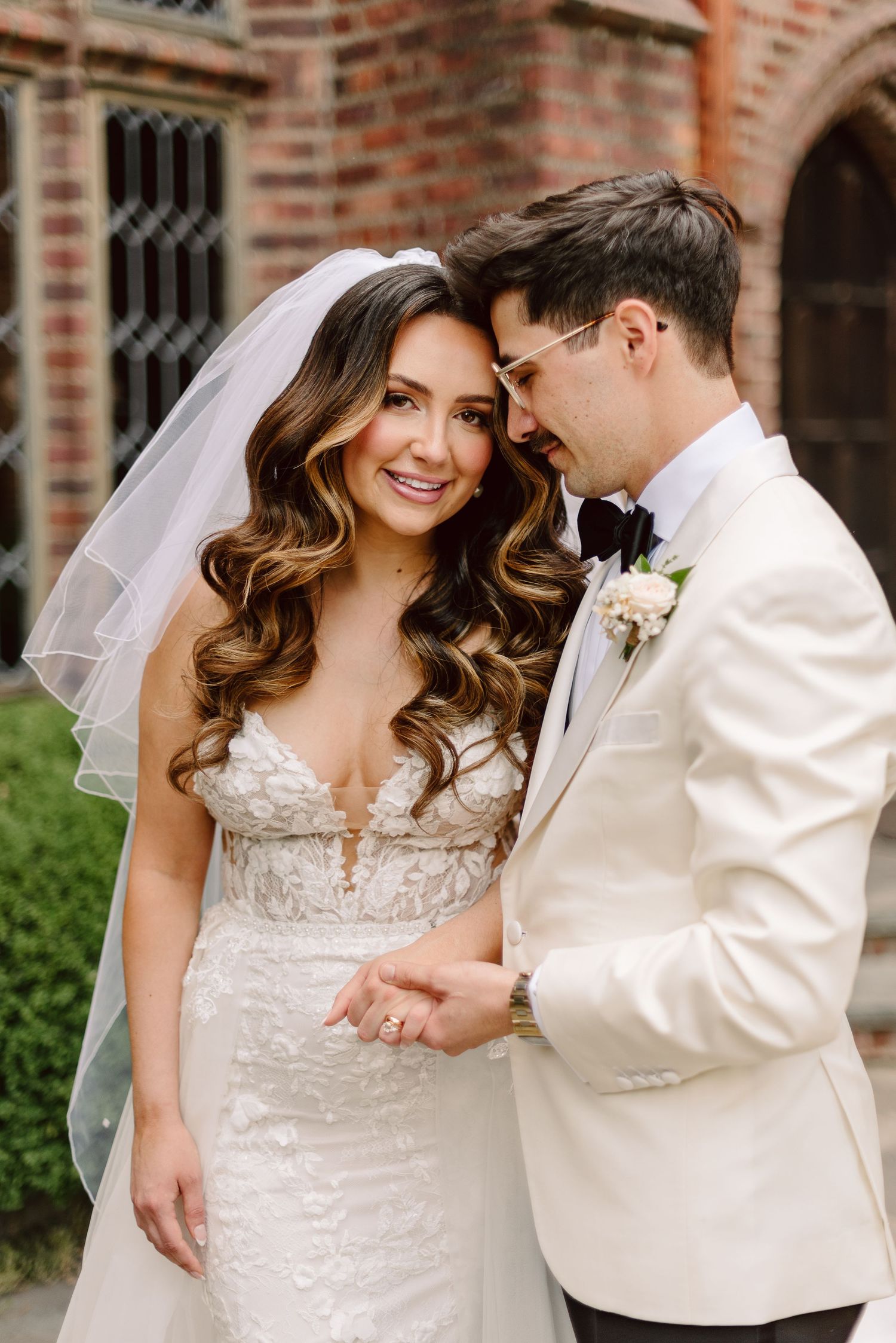 Newlywed couple in intimate moment outdoors, bride wearing lace gown with veil and groom in cream-colored suit jacket.