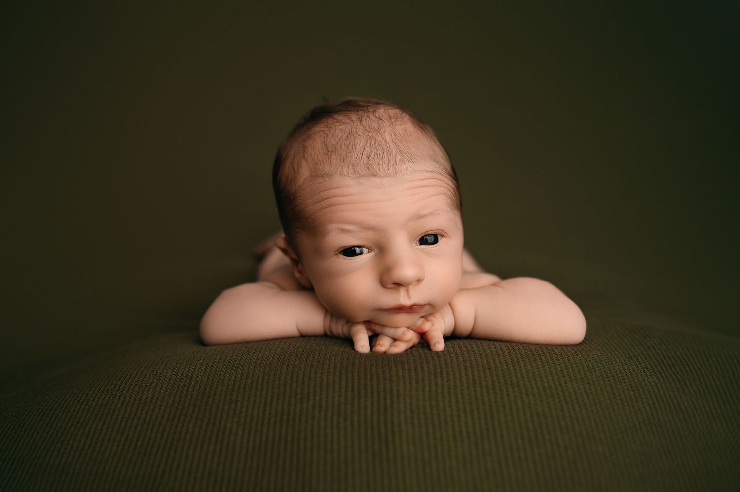 A newborn baby rests their head on crossed arms against a dark green textured background in a studio portrait.