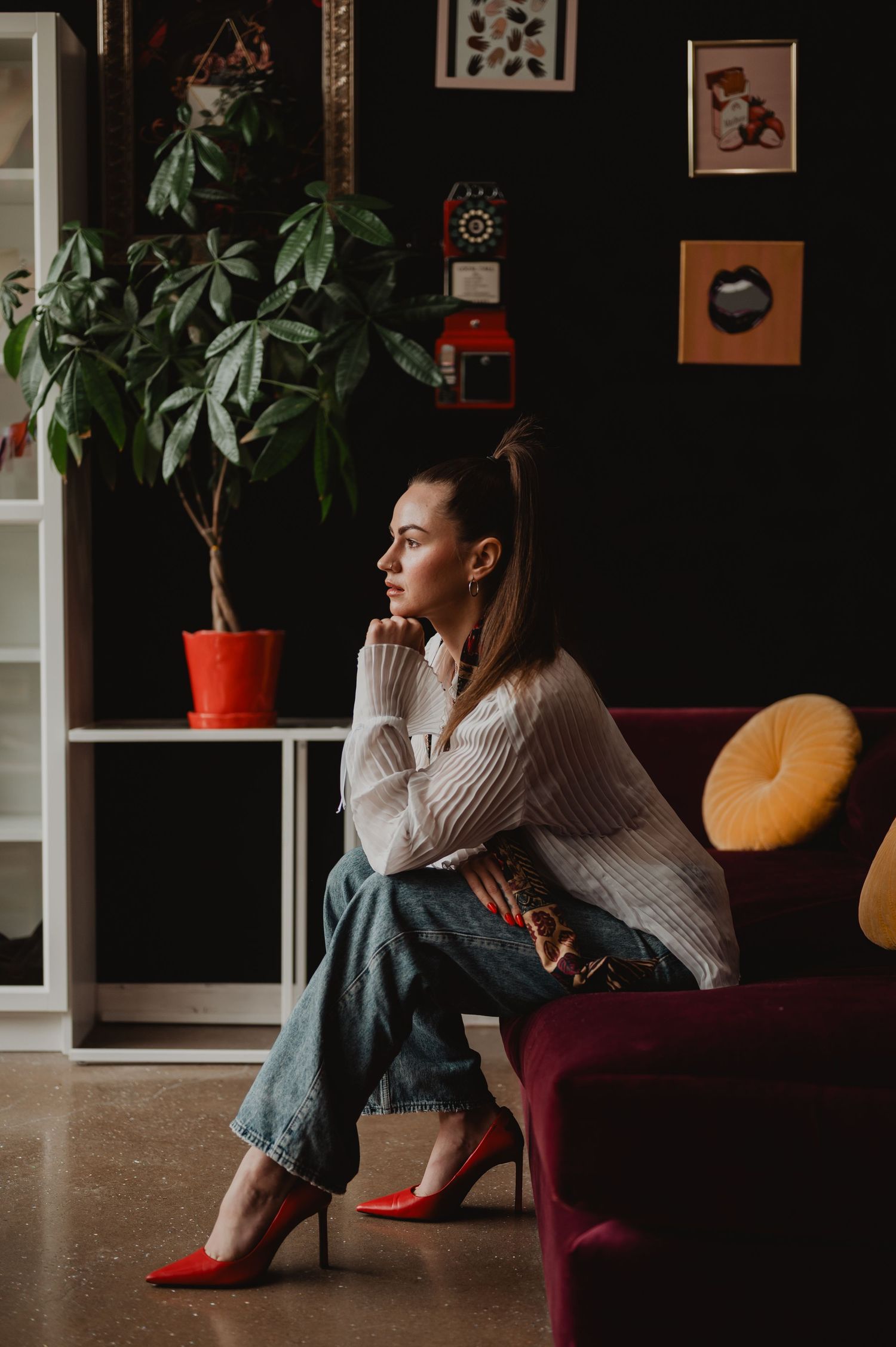 A person in a white sweater sits thoughtfully on a red chair in a cozy room with framed artwork and houseplants.
