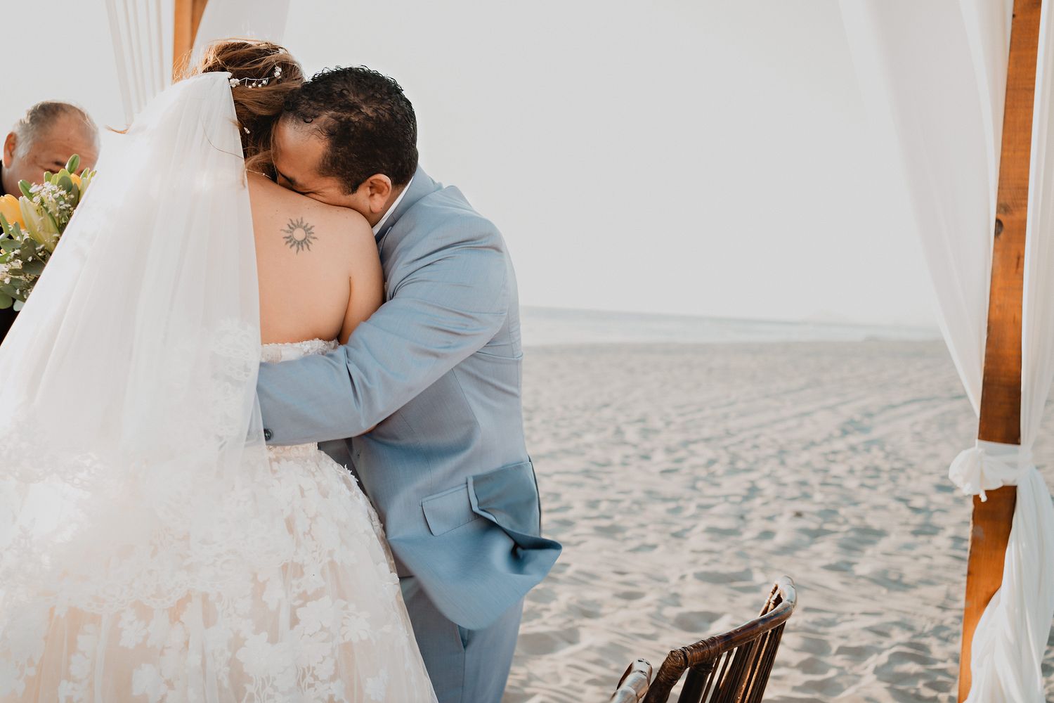 A tender embrace between newlyweds wearing light blue suit and white wedding dress on a sandy beach.