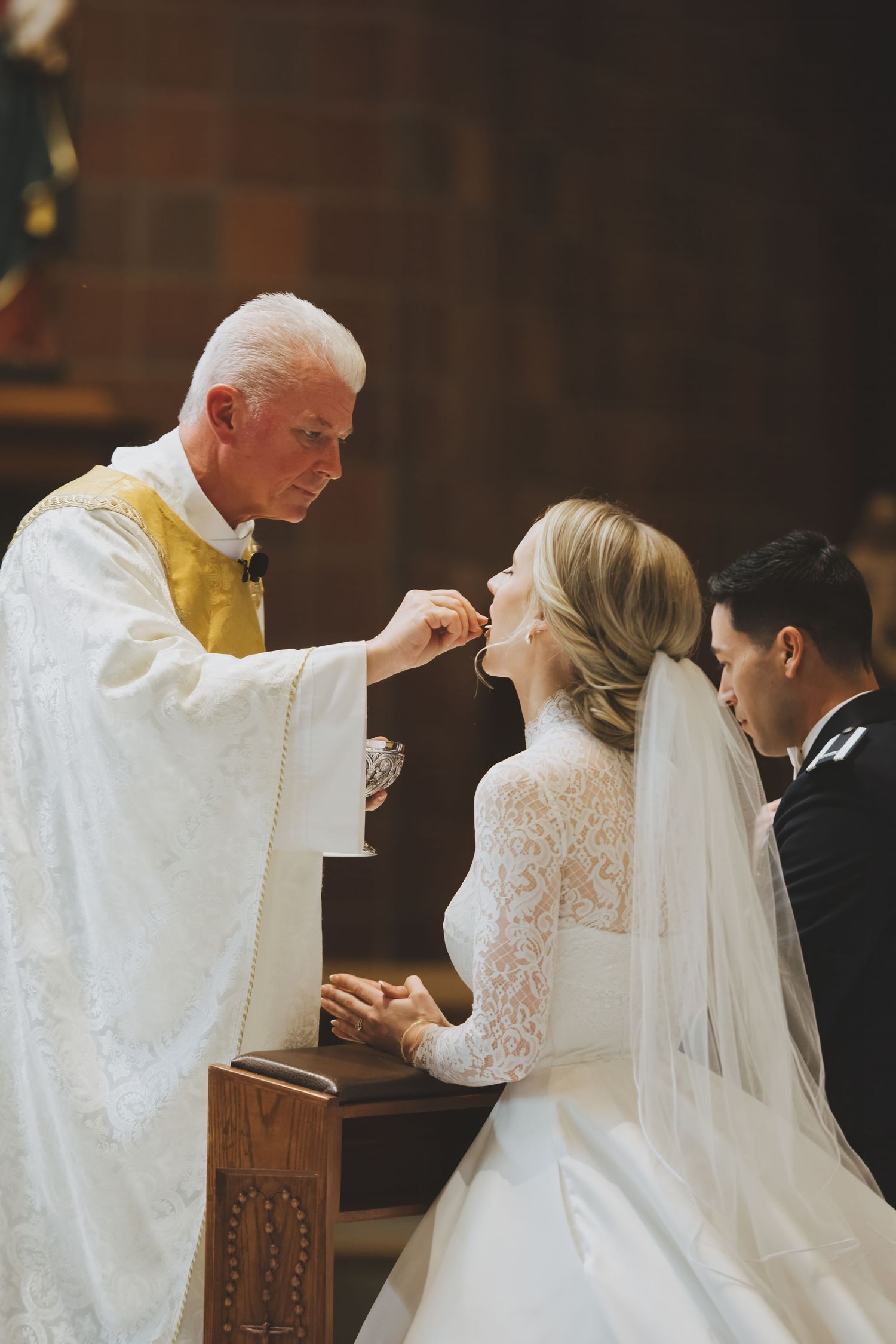 A priest administers communion during a Catholic wedding ceremony while the bride kneels in her lace gown and veil.