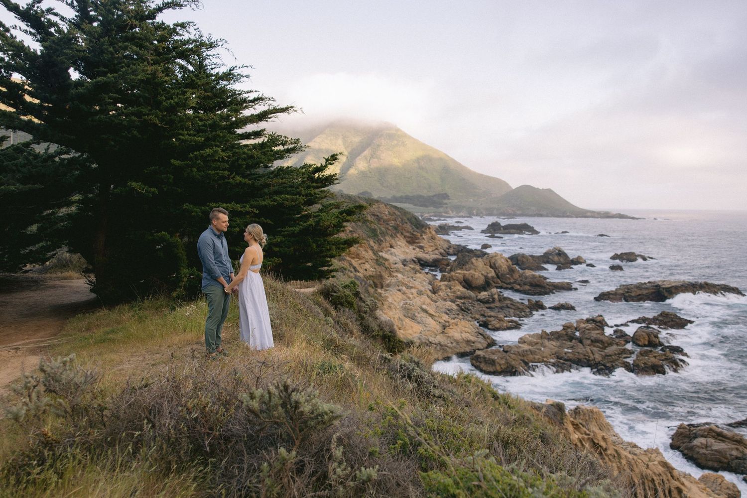 A couple stands on a scenic coastal cliff overlooking the rocky Pacific shore and ocean waves at sunset.