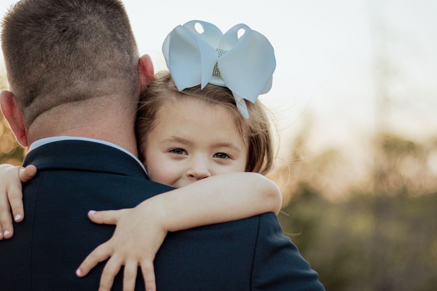 Small child wearing light blue ears hugs someone in a navy suit from behind while outdoors at sunset.