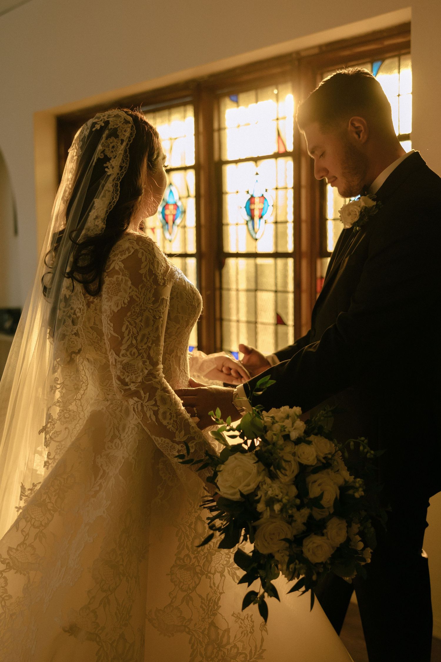 Bride and groom exchange rings at church altar bathed in golden light from stained glass windows.